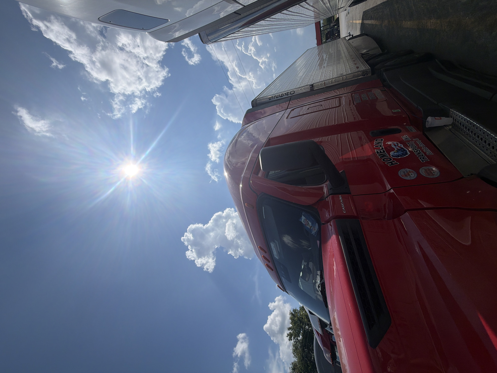 This is a low-angle, eye-level shot of the driver's side of a red semi-truck parked at a rest stop under a bright, sunny sky. The sun is visible in the upper left quadrant of the image, creating a lens flare effect. Puffy white clouds dot the bright blue sky. The truck features a white trailer, and another white truck is partially visible to the right. The truck has "HOMELAND TRANSPORT, INC." and "Buffalo Grove, IL" printed on its side, along with logos. A white disposable cup sits on the truck's running board. In the background, trees and a gas station canopy are visible. The scene is likely a truck stop or rest area, possibly in Lebanon, United States of America, given the presence of semi-trucks and the outdoor setting.