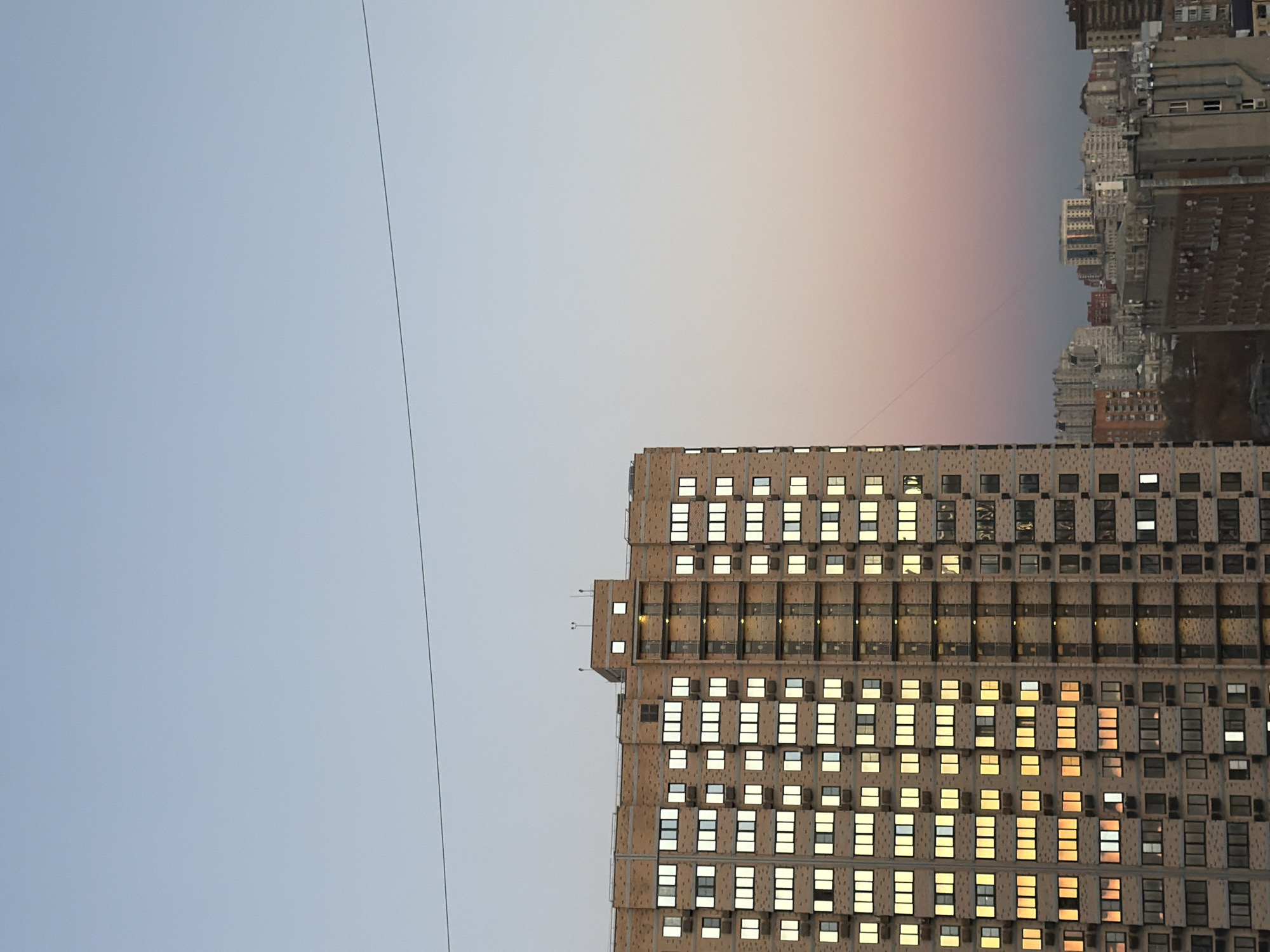 The image shows a cityscape in Rostov-on-Don, Russia, at dusk. A tall apartment building occupies the right side of the frame, its windows illuminated with warm light. The building's facade has a repetitive pattern of windows and architectural elements. To the left, the sky fades from pale blue to a soft pink hue near the horizon. In the distance, a cluster of other buildings is visible, silhouetted against the twilight sky. A thin cable runs across the upper left portion of the frame.