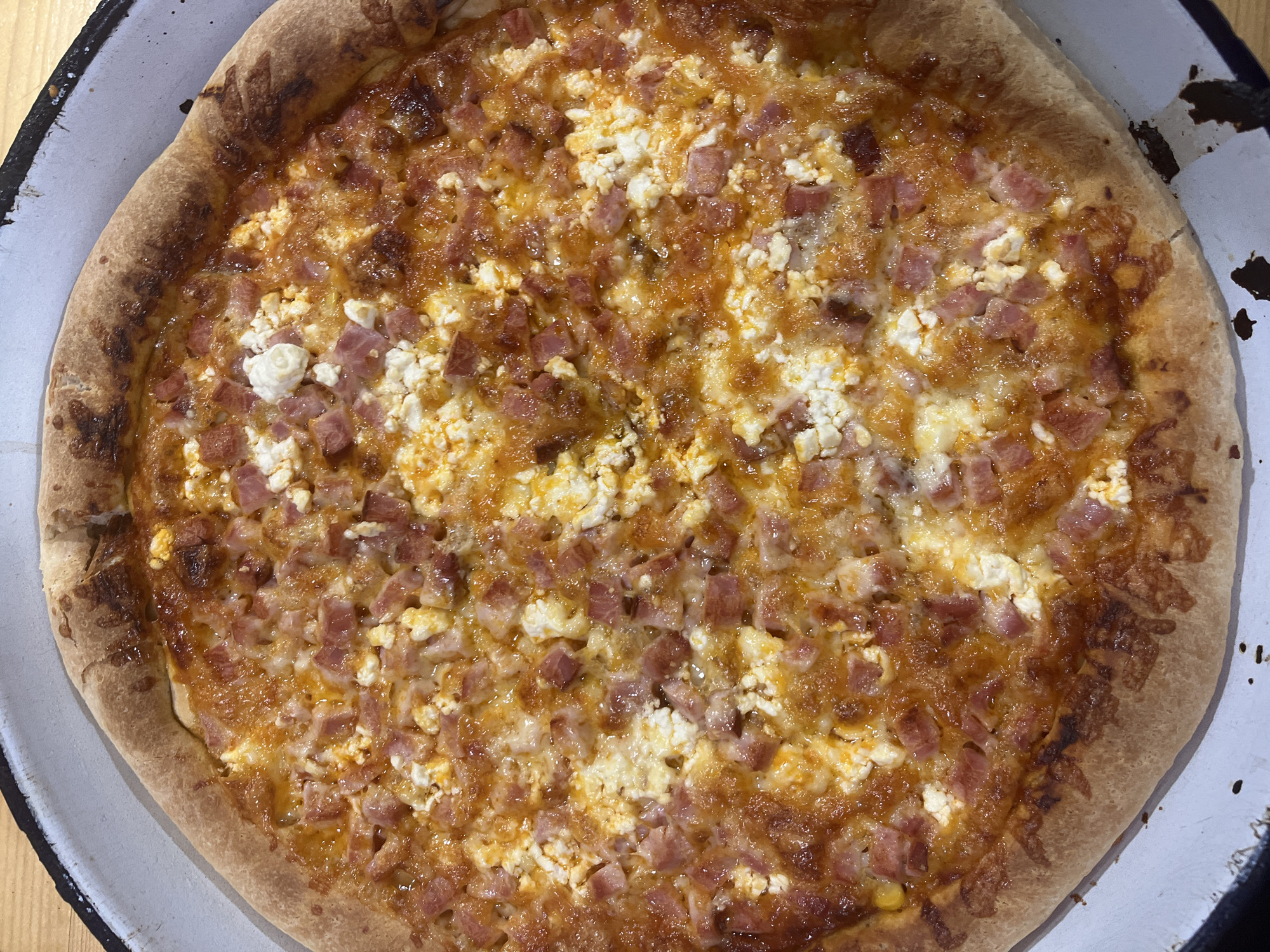 The image shows a whole pizza, possibly homemade, resting on a white enameled tray with a dark rim. The pizza has a light brown crust, a base of orange sauce, cubed ham, and scattered crumbles of white cheese. The pizza appears to be located indoors, on a wooden surface. There is no information to determine a location in Varshilo, Bulgaria.