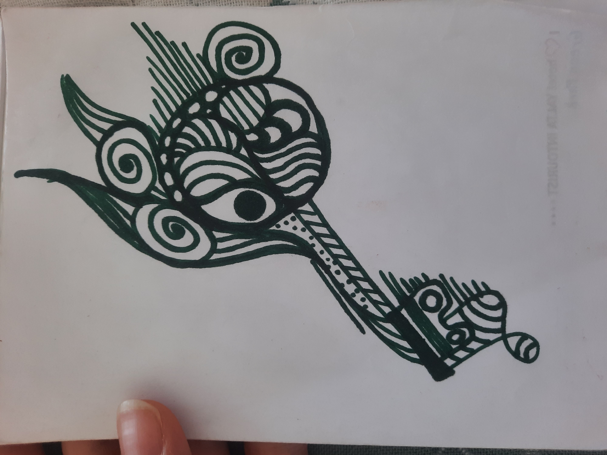 The image is a close-up, high-angle shot of a hand holding a piece of white paper with a black ink drawing on it. The drawing is of an abstract, key-like object with ornate details, including spirals, lines, and dots. The background of the image is a textured, light-colored surface. There is no discernible location in the city of Simferopol, Ukraine, or any other specific geographic context within the image.