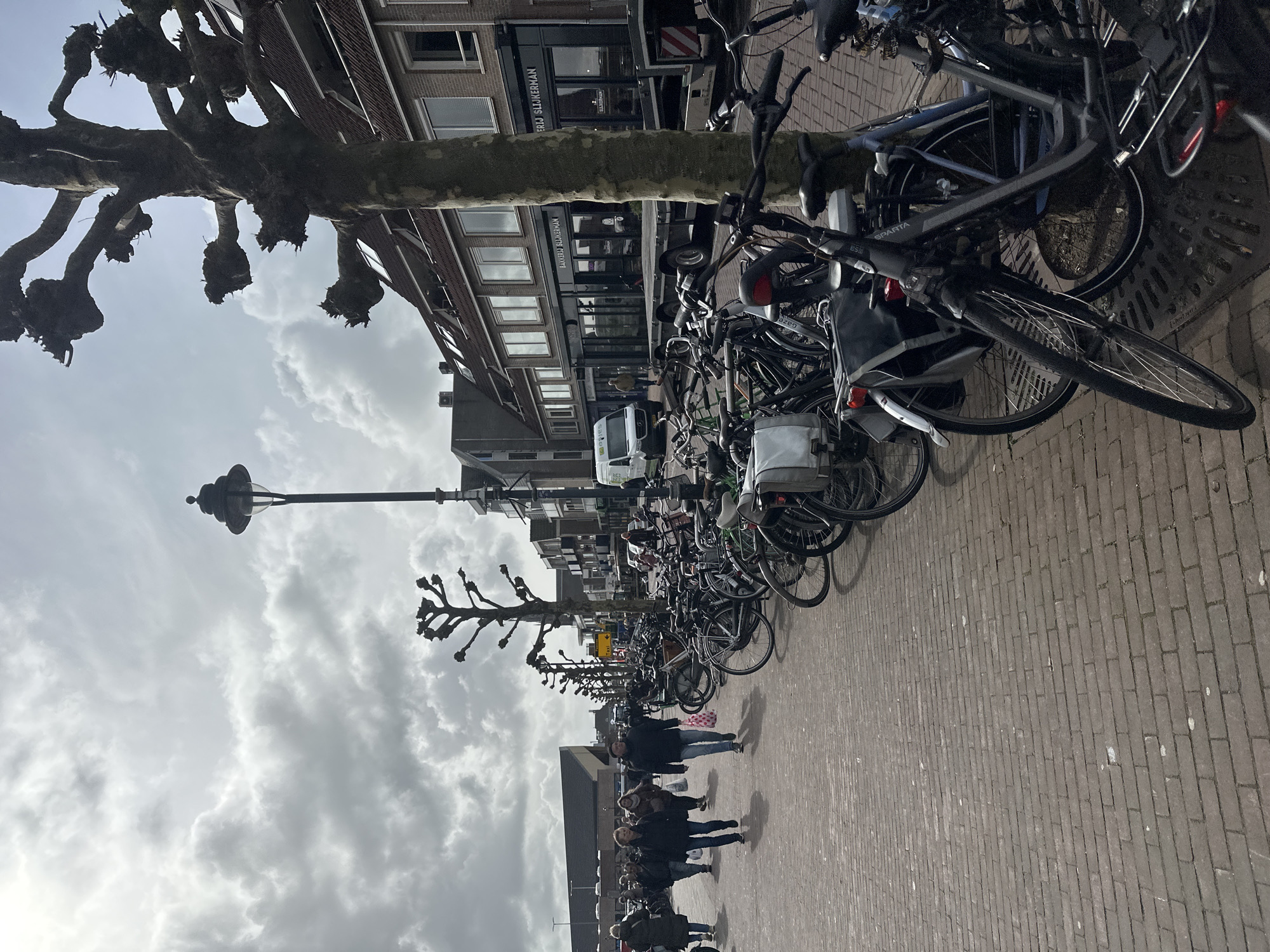 The image depicts an urban street scene in Schagen, Netherlands, under an overcast sky with diffused daylight. The perspective is from a pedestrian level looking down a street.
In the foreground, a brick-paved walkway extends from the left, narrowing into the distance. The right side of the foreground and midground is occupied by a dense, unorganized array of approximately 50-70 parked bicycles, many equipped with panniers, baskets, or child seats. A "SPARTA" branded bicycle is prominent on the right. Several large, pollarded trees with bare branches line the bicycle parking area and the walkway.
Multiple pedestrians, estimated between 10 and 15 individuals, walk along the brick pathway on the left, generally moving away from the camera. They are dressed in casual, layered clothing such as jackets and jeans; one individual carries a pink-patterned bag.
In the midground, a tall, ornate black lamppost stands upright. Beyond the parked bicycles, a two-lane street or service road is visible. A white commercial van, appearing to be a Mercedes-Benz Sprinter, is present on the street, partially obscuring a black flatbed trailer with a rear ramp. A person in a light-colored jacket stands near the trailer.
To the right, a row of two-story buildings with brick facades, large windows, and reddish-brown tiled roofs lines the street. One prominent building displays "BAKKERIJ SLIJKERMAN" on its dark storefront. Further down the street on the left, a large, dark-roofed angular building is discernible in the background. The sky is largely covered by dense, grey clouds.