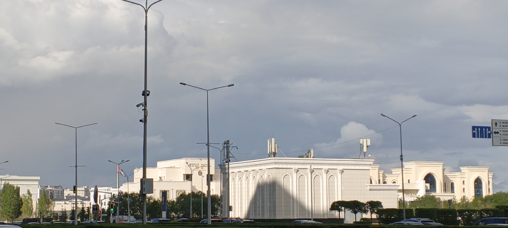 This image depicts a street scene in Astana, Kazakhstan, under a cloudy sky. The foreground shows a road with several cars. In the middle ground, there are several modern buildings with distinctive architectural features, including arched windows and decorative moldings. One building features a prominent shadow cast across its facade. Streetlights and traffic signals are visible throughout the scene. A series of road signs are present on the right side of the image, indicating directions to various locations within the city. The overall impression is of a contemporary urban environment.