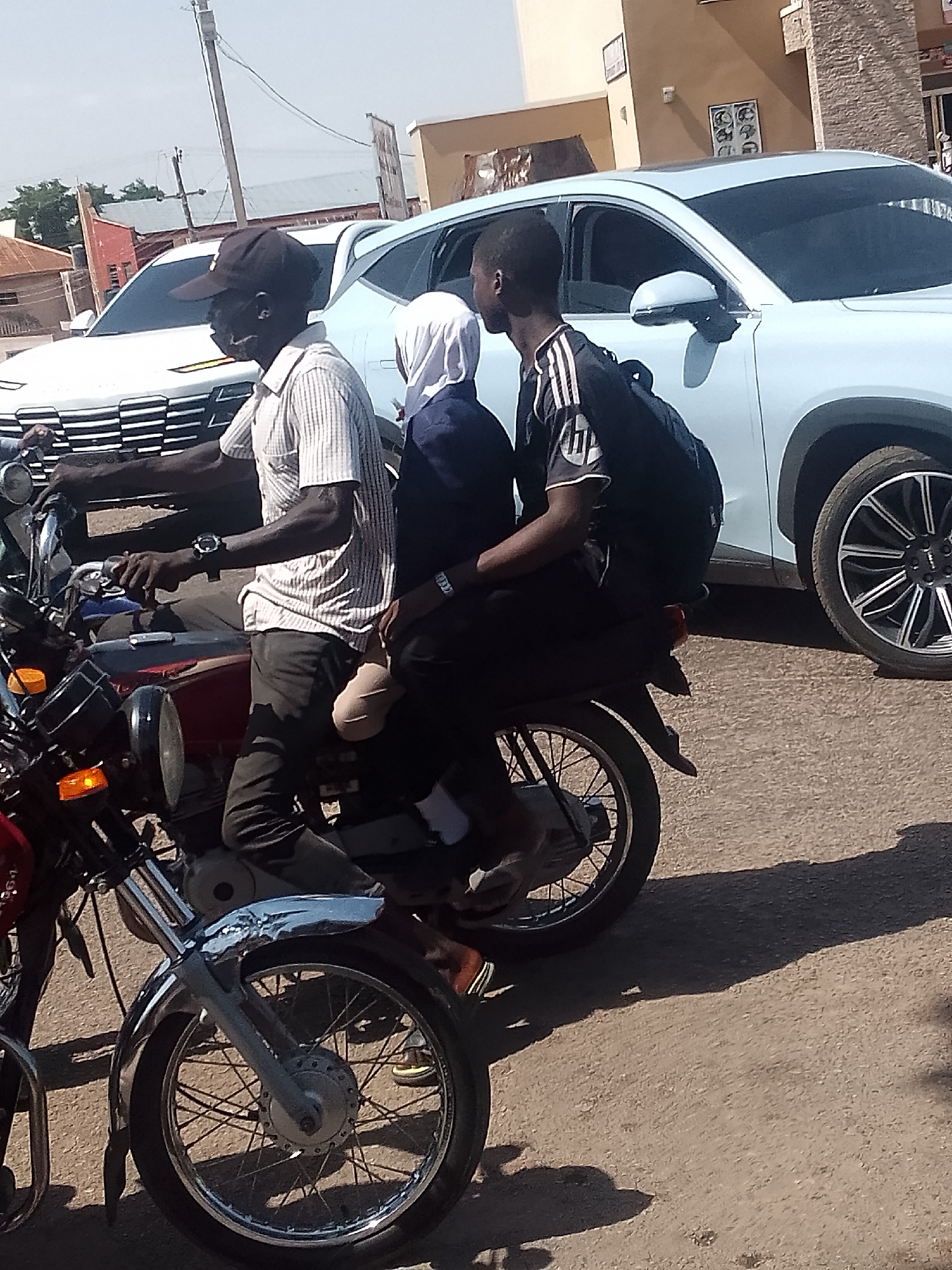 This is a daytime photograph capturing a street scene in Pantame, Nigeria, featuring three individuals on a motorcycle, stopped or moving alongside a white SUV. The lighting suggests a sunny day, with distinct shadows cast on the asphalt road.
In the foreground, a dark-skinned male driver operates a red and chrome motorcycle. He wears a black cap, a light-colored striped short-sleeved shirt, dark pants, and a watch on his left wrist. A dark face mask is pulled down to his chin. Seated directly behind him is a child or young person, possibly female, wearing a white head covering (hijab) and a dark long-sleeved top. They are positioned between the driver and the third passenger, appearing to be sitting sideways or with legs tucked due to limited space. The third passenger, also a dark-skinned male, sits at the rear, wearing a black short-sleeved shirt with white stripes on the shoulders and a prominent white "HP" logo on the chest. He carries a black backpack.
To the right of the motorcycle, a white modern SUV is visible, showing its front grill design, passenger-side door, side mirror, and part of its distinct patterned wheel. In the background, various buildings line the street, including one with a reddish roof and another with a light brown facade. A wooden utility pole with overhead wires is also visible against the bright sky. The scene depicts typical urban or semi-urban mobility in the region, with multiple passengers sharing a motorcycle.