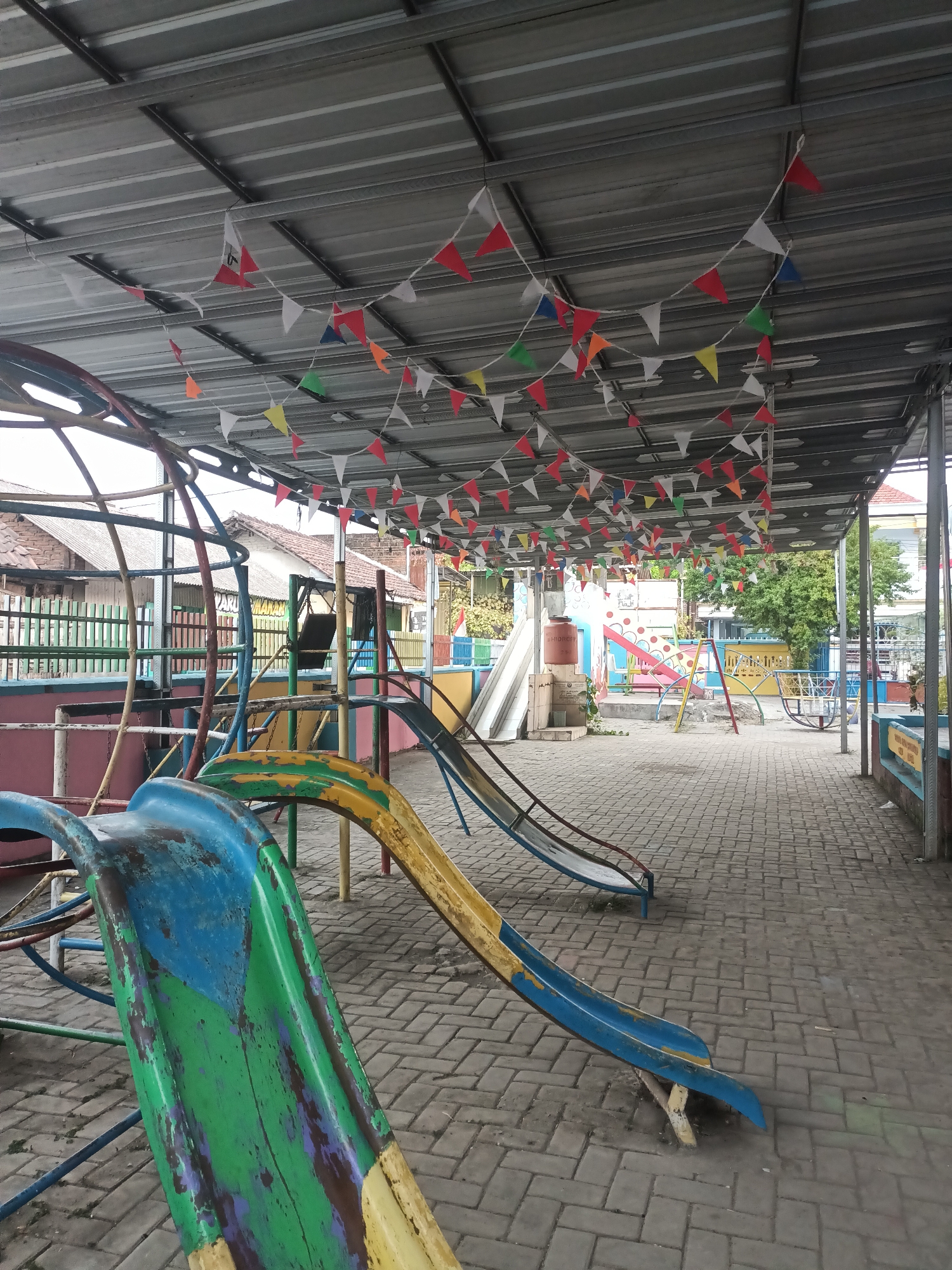 The image shows an outdoor playground area covered by a corrugated metal roof supported by metal beams. Strings of small, colorful triangular flags are strung across the underside of the roof. In the foreground, a worn, multi-colored slide, predominantly blue and green with yellow accents, is positioned diagonally. Behind it, another slide, yellow and blue, is visible. Further in the background, there are more playground structures including a set of swings and what appears to be a seesaw or spinning ride. The ground is paved with interlocking bricks. In the distance, residential buildings with tiled roofs and a colorful fence can be seen. The scene suggests a public or school playground. The location is identified as Badas, Indonesia. There are no people visible in the image.
