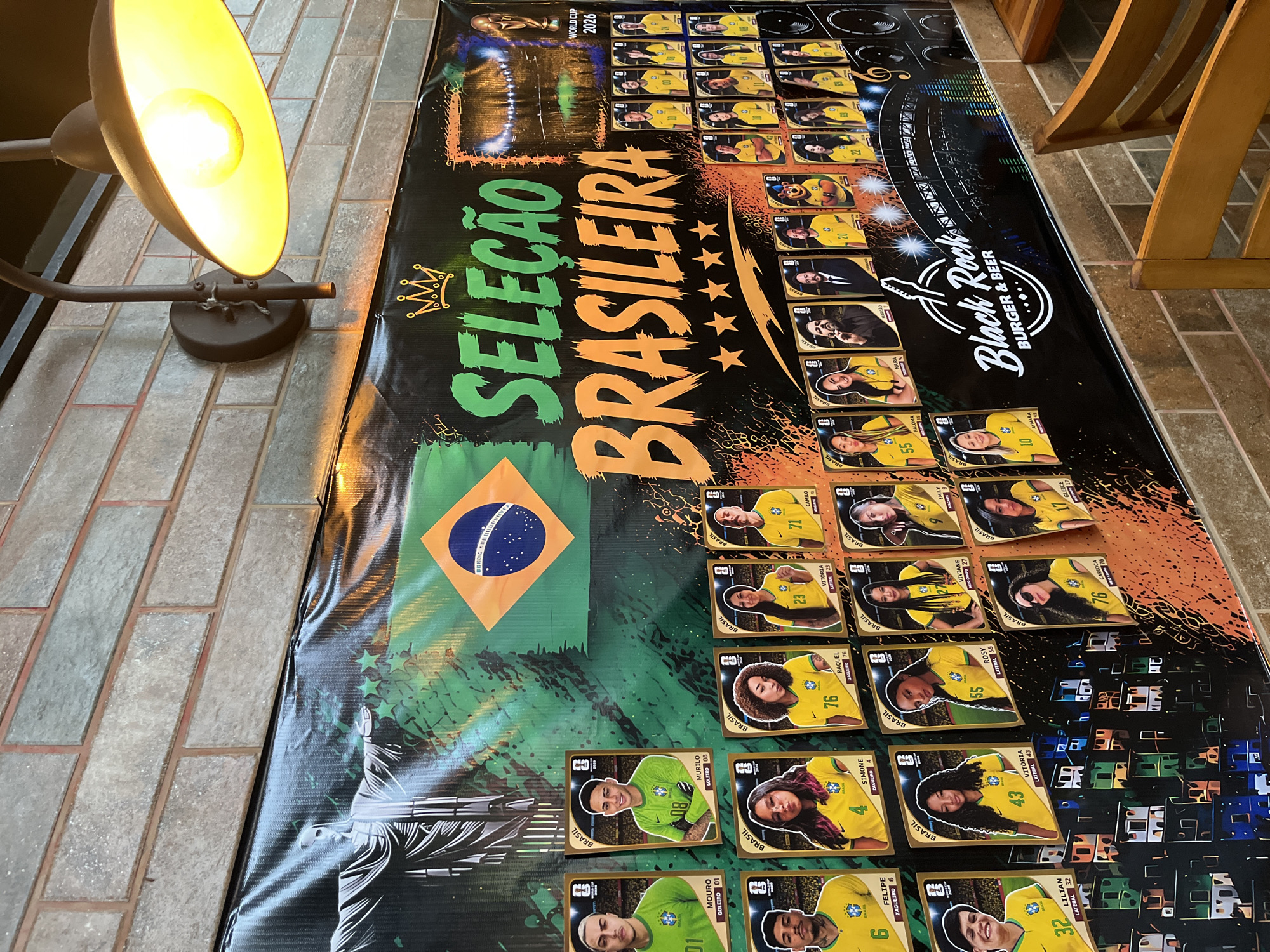 The image shows a table or surface with a banner or poster laid out on it. The poster has the Brazilian flag and the words "Seleção Brasileira," along with the phrase "World Cup 2026." The poster features images of Brazilian soccer players and the logo of "Black Rock Burger & Beer." The table is illuminated by a warm-toned lamp on the left side of the frame. The surface around the table appears to be brick or tile.