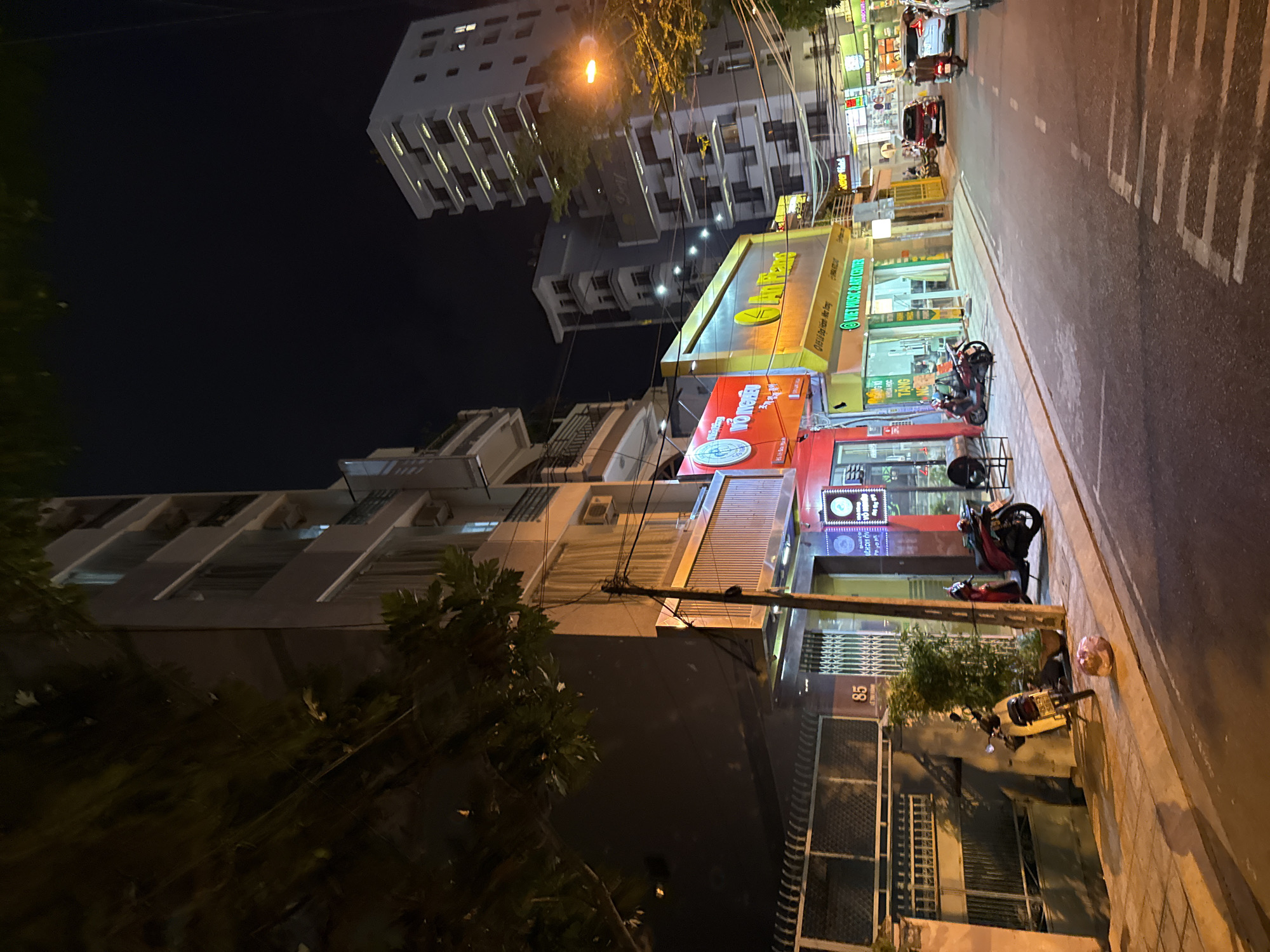 This image captures a street scene at night in Nha Trang, Vietnam, likely in a commercial or residential area. The foreground shows a paved street with white dashed lines indicating traffic lanes. A sidewalk runs along the right side of the street, lined with several businesses.
On the right side of the sidewalk, there are at least three shops with brightly lit signs. One sign is primarily yellow and features text that appears to be in Vietnamese, with the words "An Thang" clearly visible. Another adjacent sign is red and also displays Vietnamese text. A third sign, to the left of the yellow one, is green and reads "VIET MUSIC & ART CENTER". Behind these storefronts, multi-story buildings rise up, some with balconies.
On the left side of the street, a building with a dark facade is visible, with windows and balconies. A residential-looking fence with metal bars runs along the base of this building. The number "85" is visible on a pole near a gate.
Several motorcycles are parked on the sidewalk. A light-colored motorcycle is parked near the building on the left, and a red motorcycle is parked in front of one of the shops. Further down the street, there are cars and other motorcycles, suggesting ongoing activity. The sky is dark, indicating it is nighttime. Trees with dark foliage are present in the upper left portion of the image, partially obscuring the view of the buildings. Streetlights and the illuminated signs provide the primary illumination.