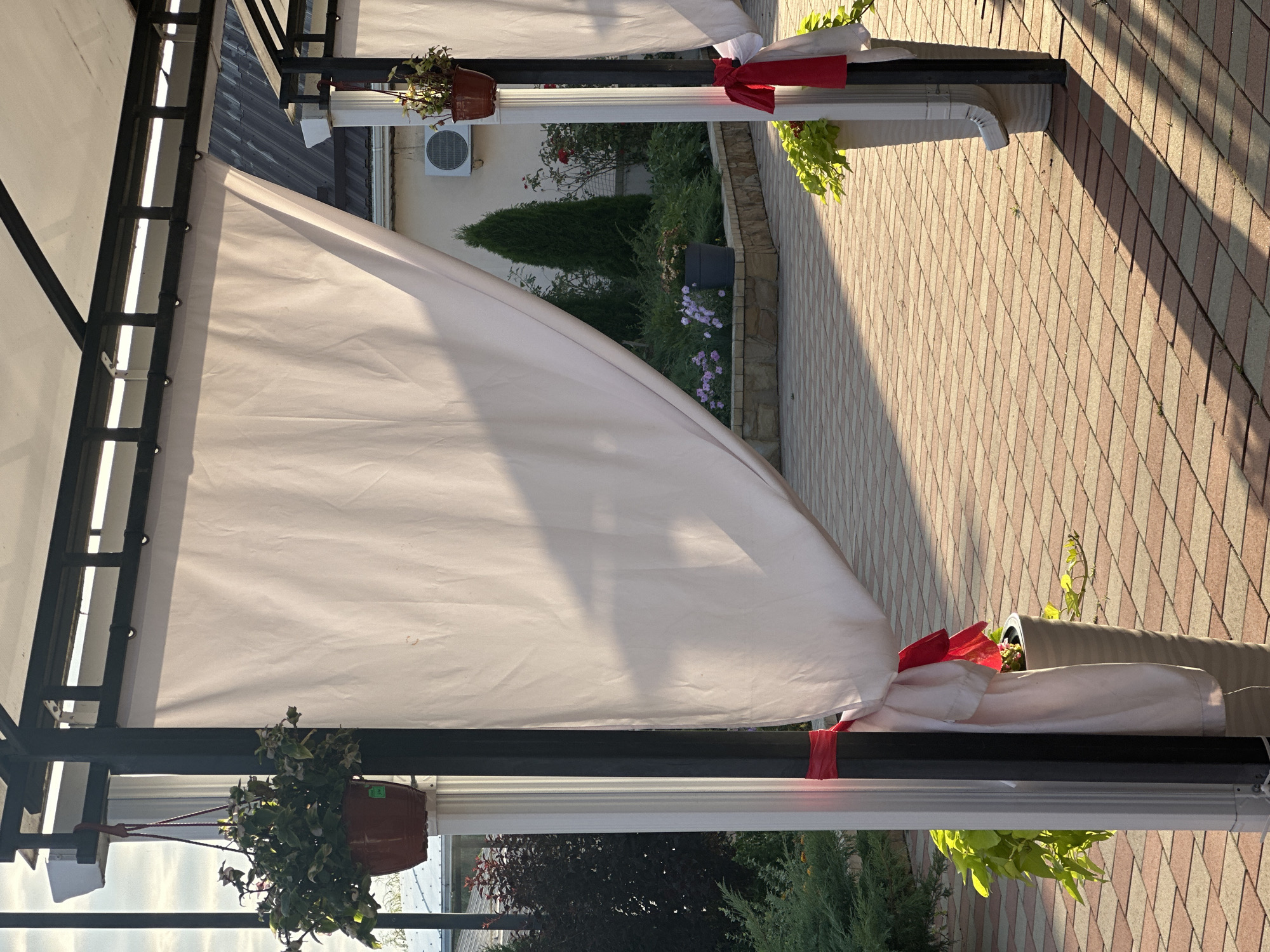 The image depicts an outdoor dining area with white drapes hanging from a black metal frame, likely part of a gazebo or awning. The drapes are tied back with red ribbons. The ground is paved with interlocking bricks in shades of red and tan. To the left, a potted plant hangs from the frame. In the background, there is a garden area with various plants, including small flowering bushes and two tall evergreen trees. A building with an air conditioning unit is visible behind the garden. The scene appears to be set in a commercial establishment or a well-maintained outdoor space, possibly a cafe or restaurant, located in Yaroslavskaya, Russia. Sunlight casts long shadows across the paved area, indicating it is either early morning or late afternoon.
