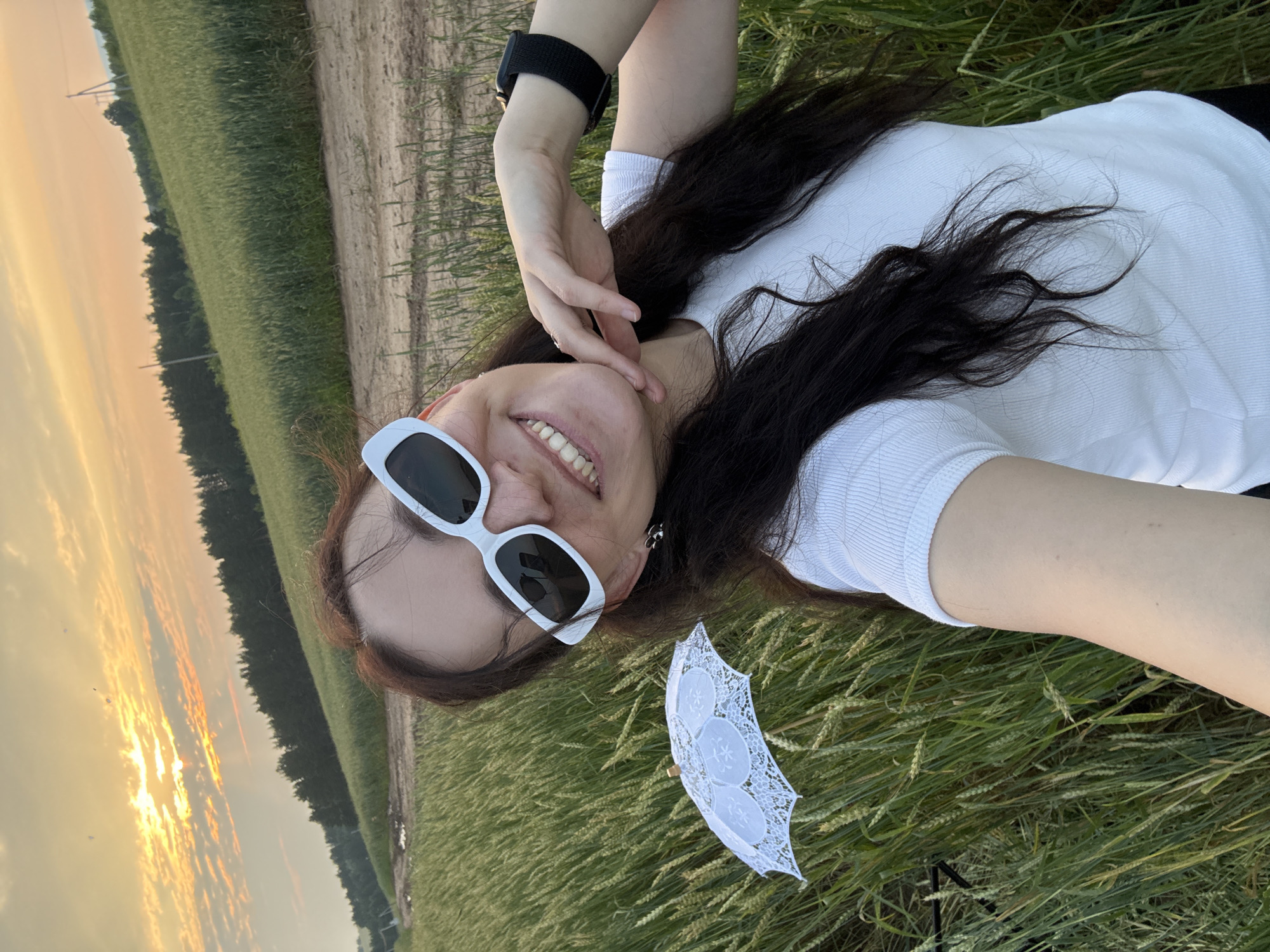 A young woman with long, dark hair is taking a selfie in a field of tall green grass. She is wearing white sunglasses and a white t-shirt. A small white lace umbrella is positioned in the grass to her left. The background shows a dirt road and a line of trees against a sunset sky. The location is the city of Fokino, Russia.