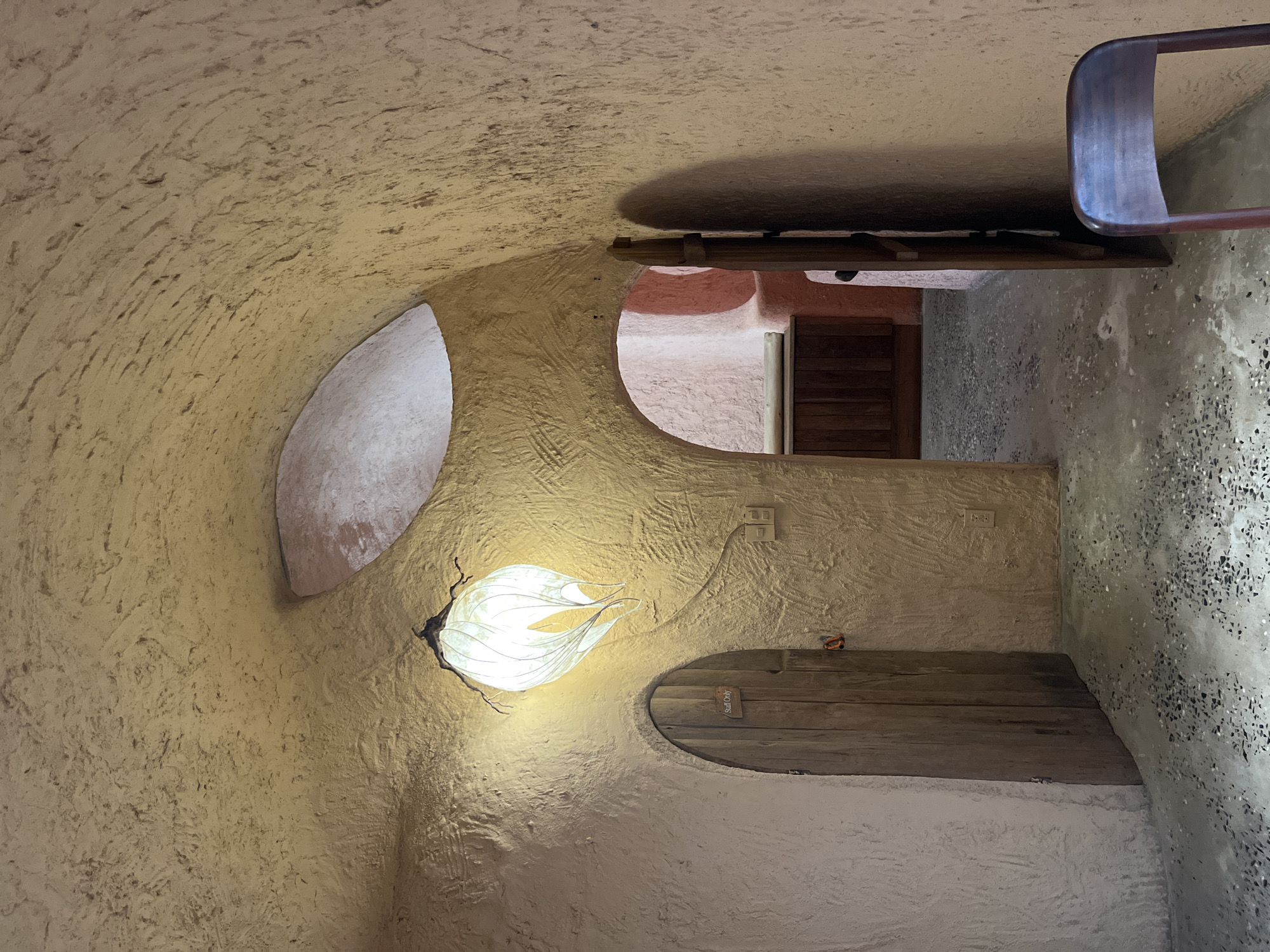 The image depicts an interior space with a curved, textured wall and ceiling. The wall appears to be constructed of a light-colored, possibly adobe-like material. There is a decorative light fixture resembling a white flower mounted on the wall. The room has an opening to another space beyond, where a wooden door is visible. Another opening in the wall is present, and below it, a wooden ledge with a sign that says "Staff Only" is installed. There is an electrical outlet visible near the ledge. A partial view of a chair is visible at the top of the frame. The location is listed as Unknown, Unknown.