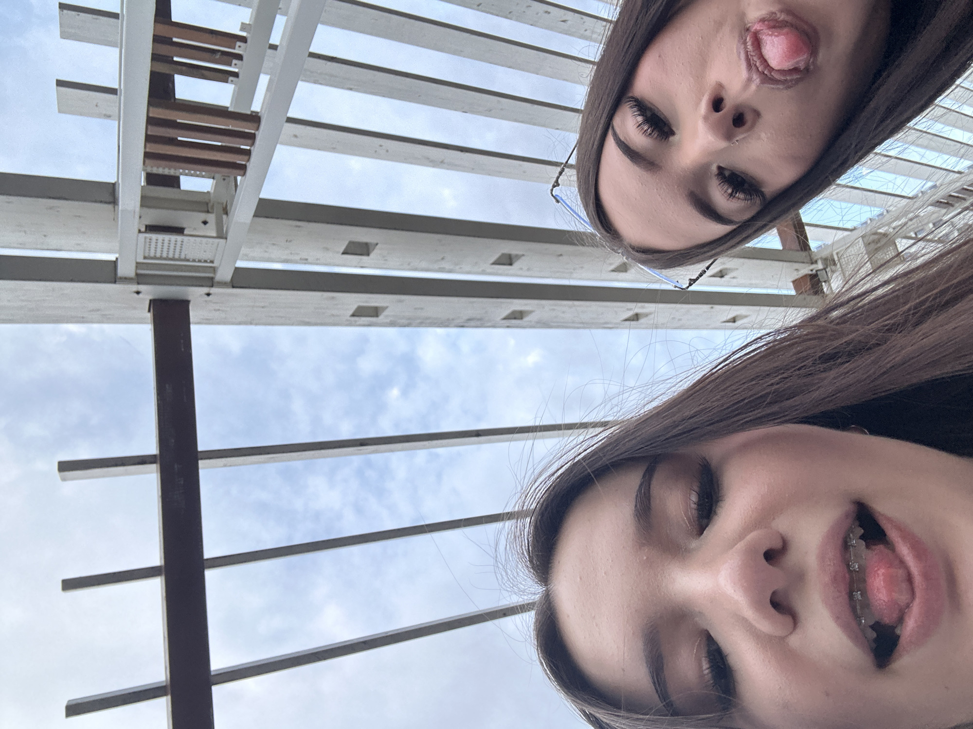 Two young females are captured in a low-angle selfie, positioned in the foreground. The individual on the left is closer to the camera and has her mouth open, revealing braces on her teeth, and her tongue is protruding. The individual on the right is slightly further back, also with her mouth open and tongue sticking out. She is wearing glasses that rest on her nose. Both subjects have dark hair and appear to be outdoors.
The background consists of a structure made of light-colored beams, possibly a pergola or similar architectural element, against a sky with visible clouds. The perspective suggests the photo was taken from below, looking upwards at the subjects and the sky. The scene takes place outdoors in an urban environment, likely Naberezhnye Chelny, Russia, given the context.