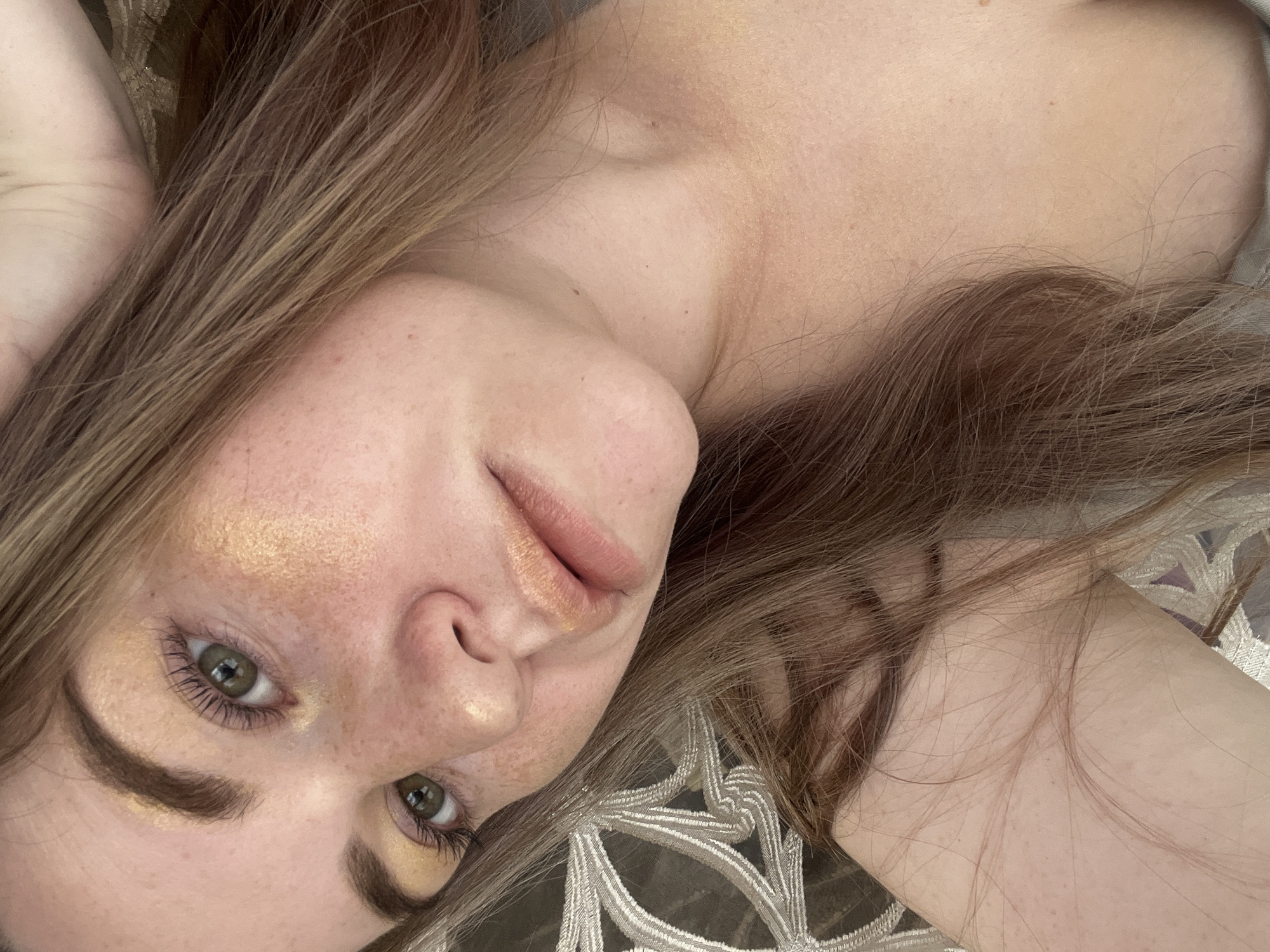 A close-up image depicts a fair-skinned female individual, appearing to be a young adult, captured in an upward-angled shot. Her long, light brown hair frames her face and extends over her visible shoulders and upper chest. Distinctive features include green eyes with long, dark eyelashes, defined eyebrows, and noticeable freckles across her nose and cheeks. Her makeup consists of shimmering gold or bronze tones applied to her eyelids, inner eye corners, and upper cheekbones, complementing a glossy, peachy-gold lip color. The individual's shoulders and upper chest are bare, suggesting minimal clothing or a wide-necked garment. The background is out of focus, revealing a patterned textile, possibly a pillow or blanket, featuring a light-colored, intricate, curvilinear design against a darker base, consistent with an indoor setting. The specific location within the city of Chaikovsky, Russia, cannot be determined from the visual information present in the image.