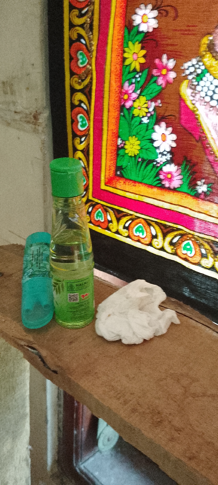 A close-up shot shows a wooden shelf with three items. On the left is a turquoise plastic bottle, next to it a clear glass bottle with a green cap containing a light yellow liquid, and to the right is a crumpled white tissue. Behind the shelf, a framed artwork with floral motifs and a dark border is visible. The background wall is textured and light-colored.