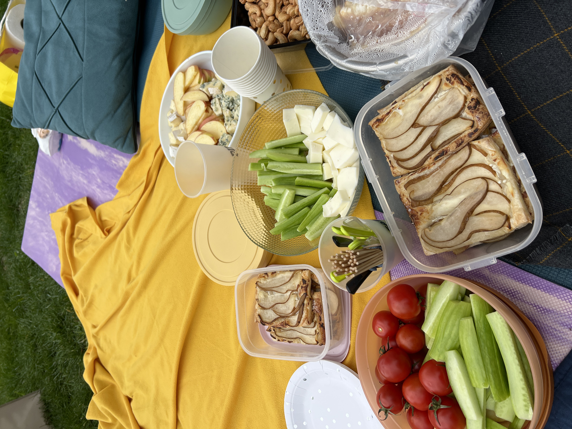 A picnic is laid out on a yellow and purple blanket on grass. There is a bowl of sliced apples and blue cheese, a stack of white paper cups, a bowl with celery sticks and cheese cubes, a container with pear tarts, a container of nuts, a container with what looks like bread, a bowl with tomatoes and celery sticks, a container of toothpicks, and some plates. There is also a blue-green pillow. The image does not clearly show any aspect that would allow a determination of whether the picnic is in the city of Tbilisi, Georgia.