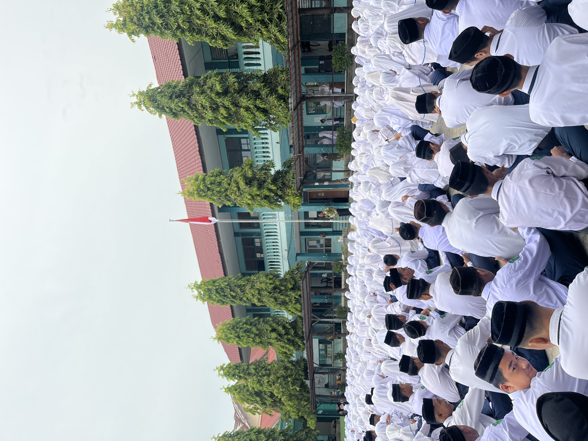 A large gathering of students, predominantly male and dressed in white shirts and dark pants with black peci hats, are seated outdoors on a paved ground. The students are facing away from the camera, with some looking towards the front of the gathering. Behind the students is a light blue building with a red roof, multiple windows, and balconies. Several tall, slender green trees are planted in front of the building. A flagpole with the Indonesian flag is visible on the left side of the building. The sky is overcast and gray. The scene appears to be an outdoor assembly or event at an educational institution. The presence of the Indonesian flag and the attire of the students suggest a formal occasion. The location is Lhokseumawe, Indonesia, likely at a school given the building and the student assembly.