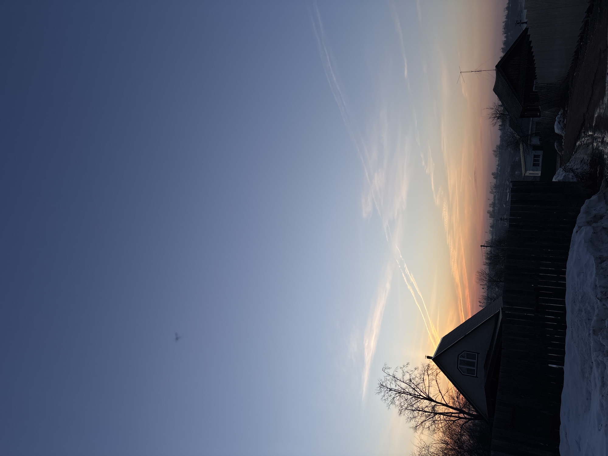 The image depicts a twilight scene in Фоки, Russia. The sky transitions from deep blue to orange, with contrails visible. Silhouetted houses, fences, and trees stand against the colorful sky. Snow is on the ground.