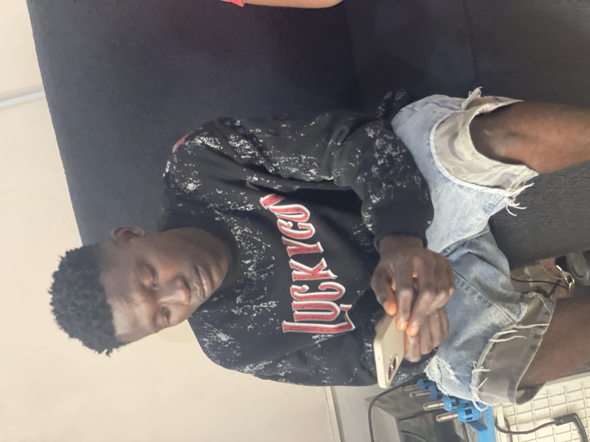 A person of African descent is sitting on a black couch, angled to the left. The individual is wearing a black sweatshirt with the word "LUCKYCOR" in red and white letters and ripped denim shorts. They are holding a smartphone in their hands. The environment appears to be indoors, with glimpses of a white wall and some metallic and blue objects near the bottom of the frame. The location is Ado, Nigeria.