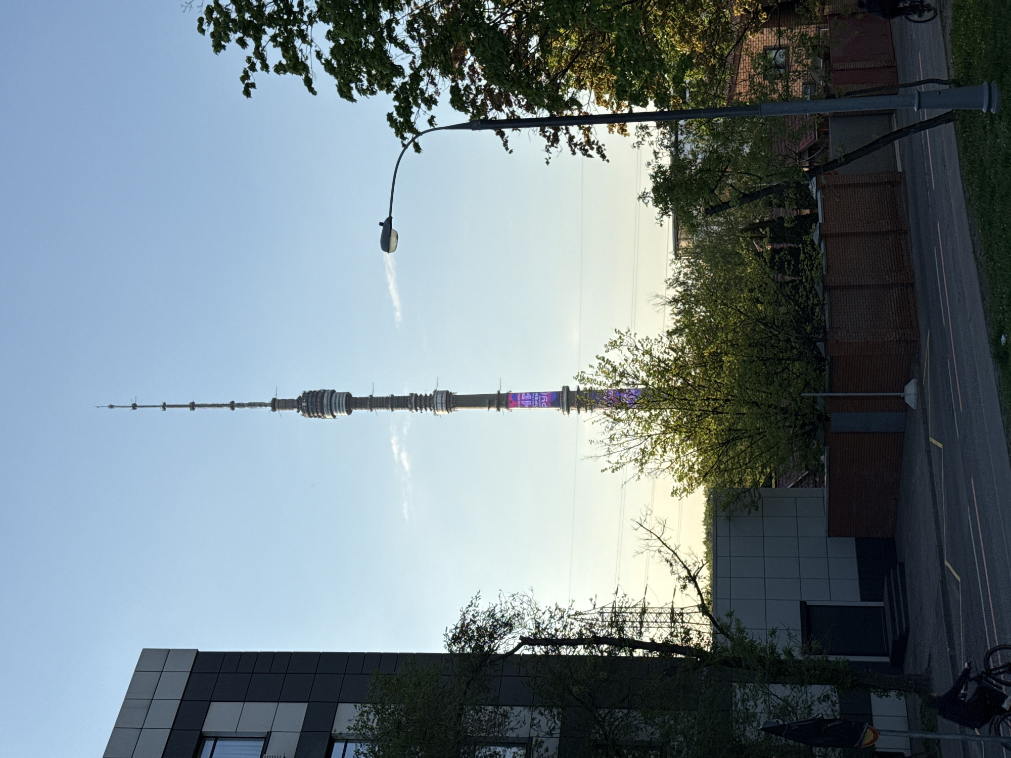 The image shows the Ostankino Tower in Moscow, Russia. The tower is tall and slender, rising into a clear, blue sky. A section of the tower is illuminated with purple lights. The surrounding urban landscape includes buildings with modern architecture, trees with green foliage, and street infrastructure such as a lamppost and a road. A brick building is visible in the upper right. Bicycles are parked near the lower right corner of the frame. The photo appears to have been taken at ground level.