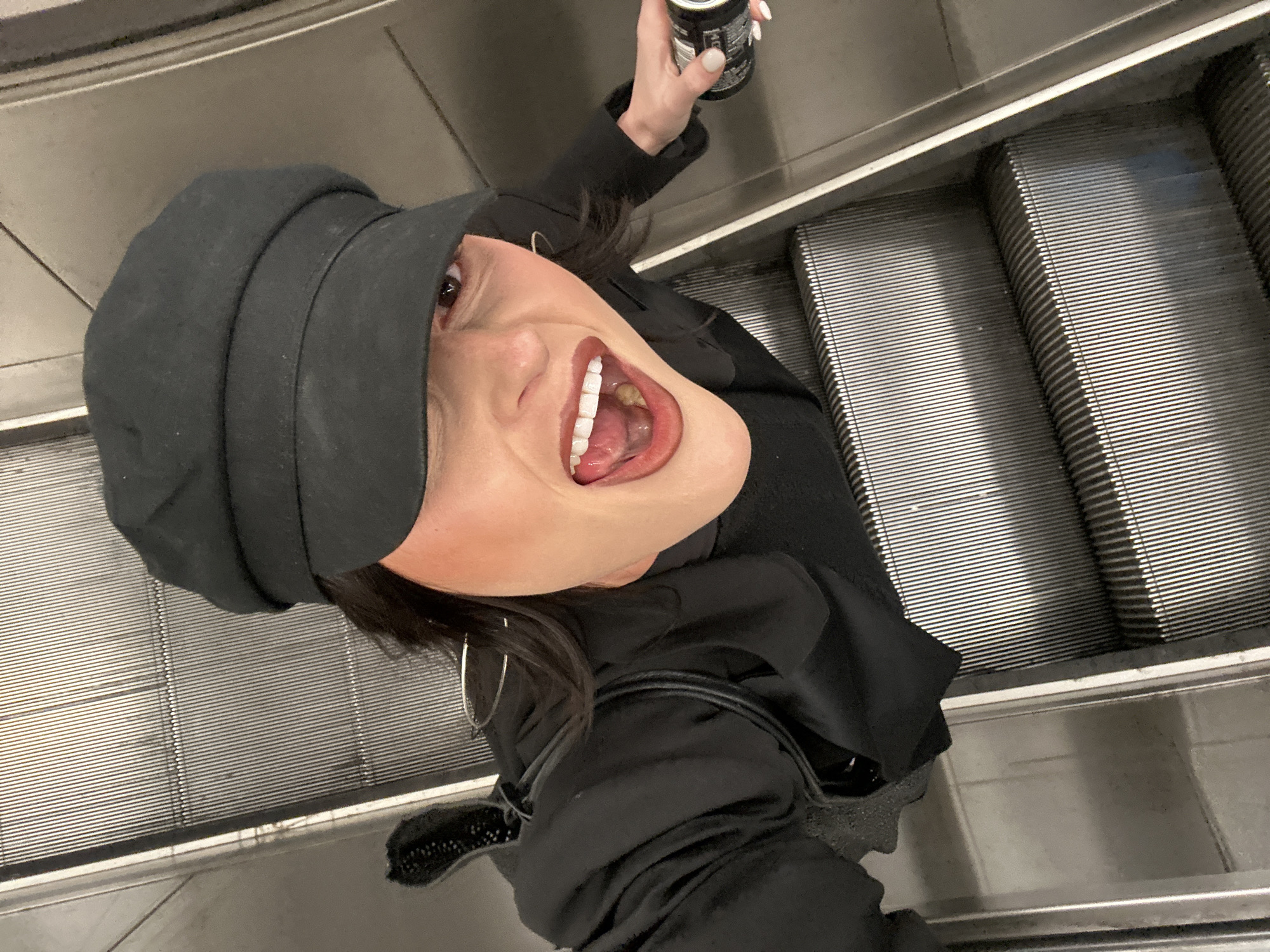 An individual, identified as female, is depicted from an elevated viewpoint on a metallic escalator within the London Underground system, London, United Kingdom. She wears a black newsboy cap and a black long-sleeved top. Her dark hair is visible beneath the cap. Her face presents an open-mouthed expression, revealing white teeth and a partially visible tongue; her eyes are open, with her right eye partly obscured by the cap brim. She has reddish-brown lipstick and visible blush. In her left hand, adorned with white-polished fingernails, she holds a black and silver beverage can. The escalator steps are composed of ribbed metallic panels. The scene captures the individual in a seemingly expressive or joyful pose, looking directly upwards towards the camera.