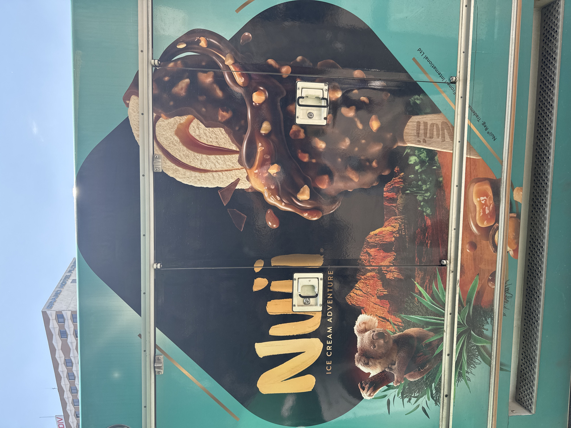 The image shows the side of a Nuii ice cream truck in Plovdiv, Bulgaria. The truck is painted with a large advertisement for Nuii ice cream, featuring an image of a chocolate and nut-covered ice cream bar, a koala, and text including "Nuii Ice Cream Adventure." A building, possibly Grand Hotel Plovdiv, is visible in the background. The image is vertically oriented.