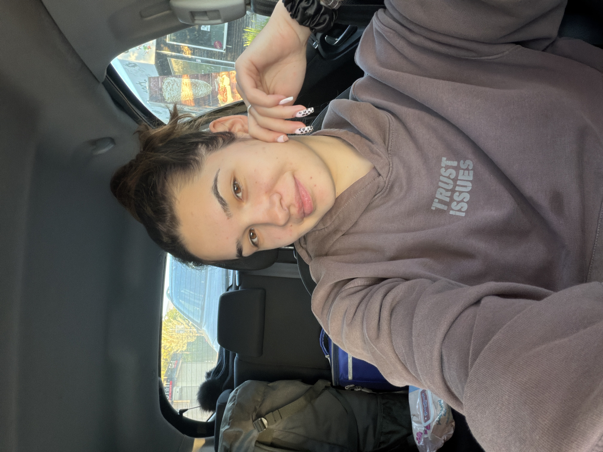 A woman is in the back seat of a car. She is fair-skinned with brown hair pulled back and is wearing a brown hoodie with the words "TRUST ISSUES" printed on it. Her nails are painted white with a black and white Dalmatian print. A backpack and a blue lunch bag are on the seat next to her. The car is parked near a commercial area. The reflection in the window shows street signs and buildings, suggesting the location is within a city. It is not possible to confirm the location in Sofia, Bulgaria.