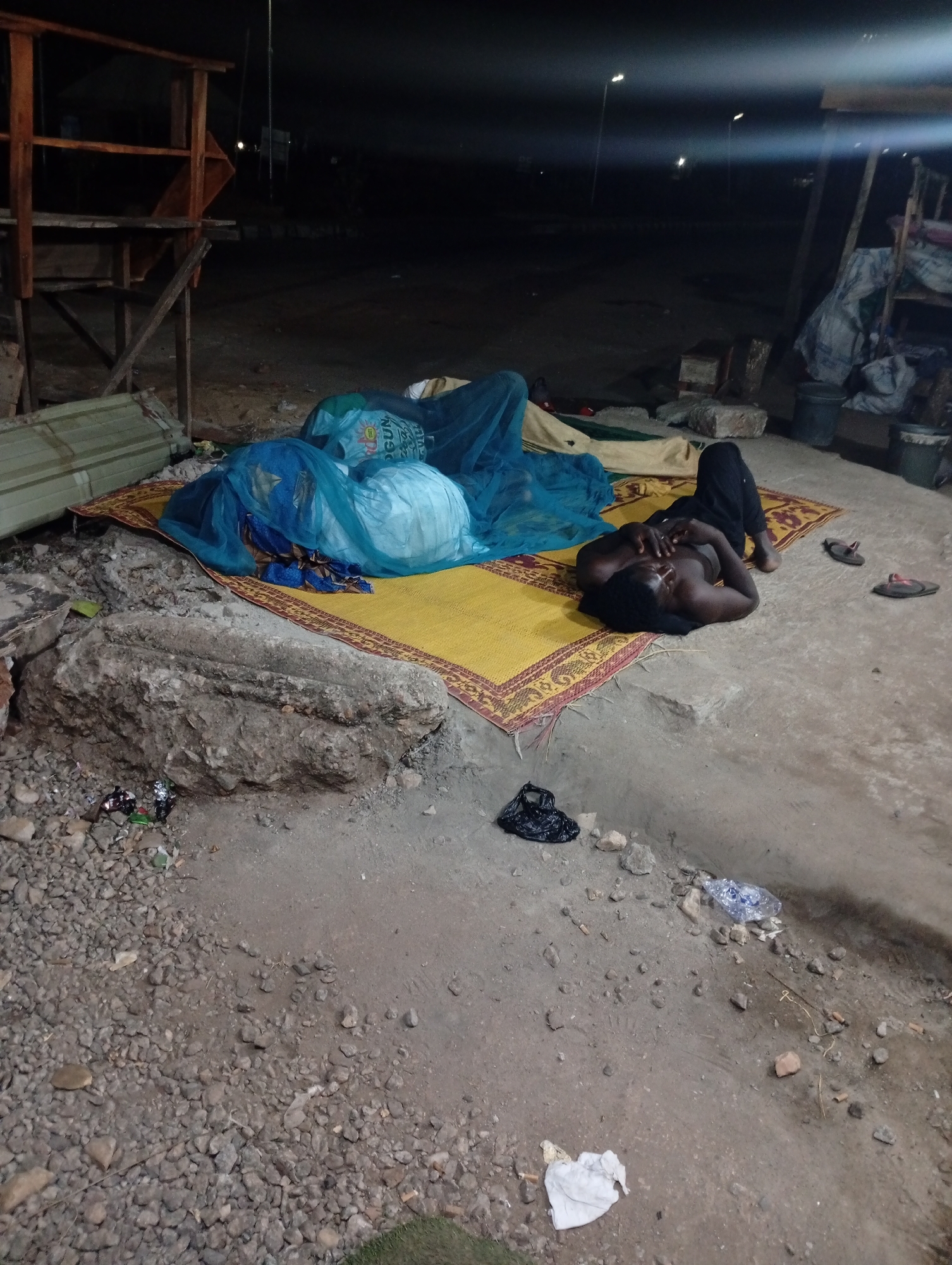 The image depicts an outdoor scene in Adankolo, Nigeria, at night. A shirtless man is lying on a yellow and red patterned mat, dressed in black pants. Next to him are two pairs of sandals. Adjacent to the mat is a large rock and scattered litter. A makeshift wooden structure is visible on the left side of the image, and another structure is on the right. Bags covered by blue netting sit on the mat. Streetlights illuminate the dark background. The ground is dirt and gravel.