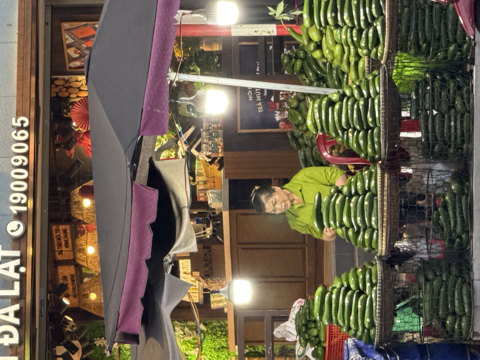 The image shows a street scene in Da Lat, Vietnam. A woman in a light green shirt stands next to a stack of cucumbers that are arranged in vertical rows inside wire baskets. A black sign behind the cucumbers displays a menu written in Vietnamese. The store front is decorated with bamboo lamps and wooden boxes, and the street side is marked by a sign with the city name "ĐÀ LẠT" and a phone number "19009065". An umbrella is open in front of the storefront.