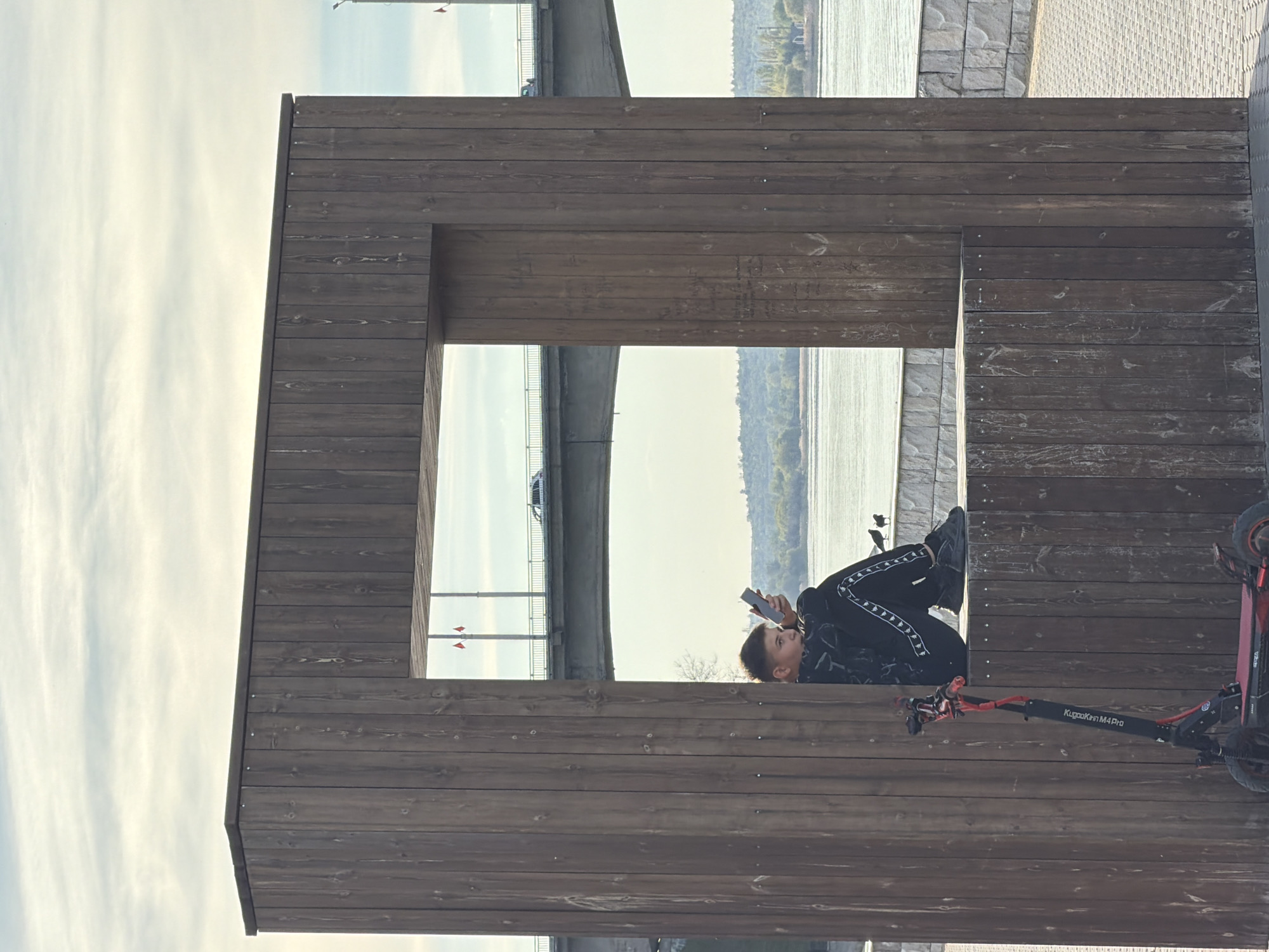 The photograph shows a young person sitting on a wooden structure, possibly a viewing platform or architectural feature, in Kamyshin, Russia. They are looking at a mobile phone. Part of an electric scooter is in the scene. The wooden structure frames a view of a river, a bridge, and the opposite riverbank. The sky is visible through another opening in the structure. The person is wearing black clothing and footwear. The scooter is red and black. The setting appears to be a public outdoor space.
