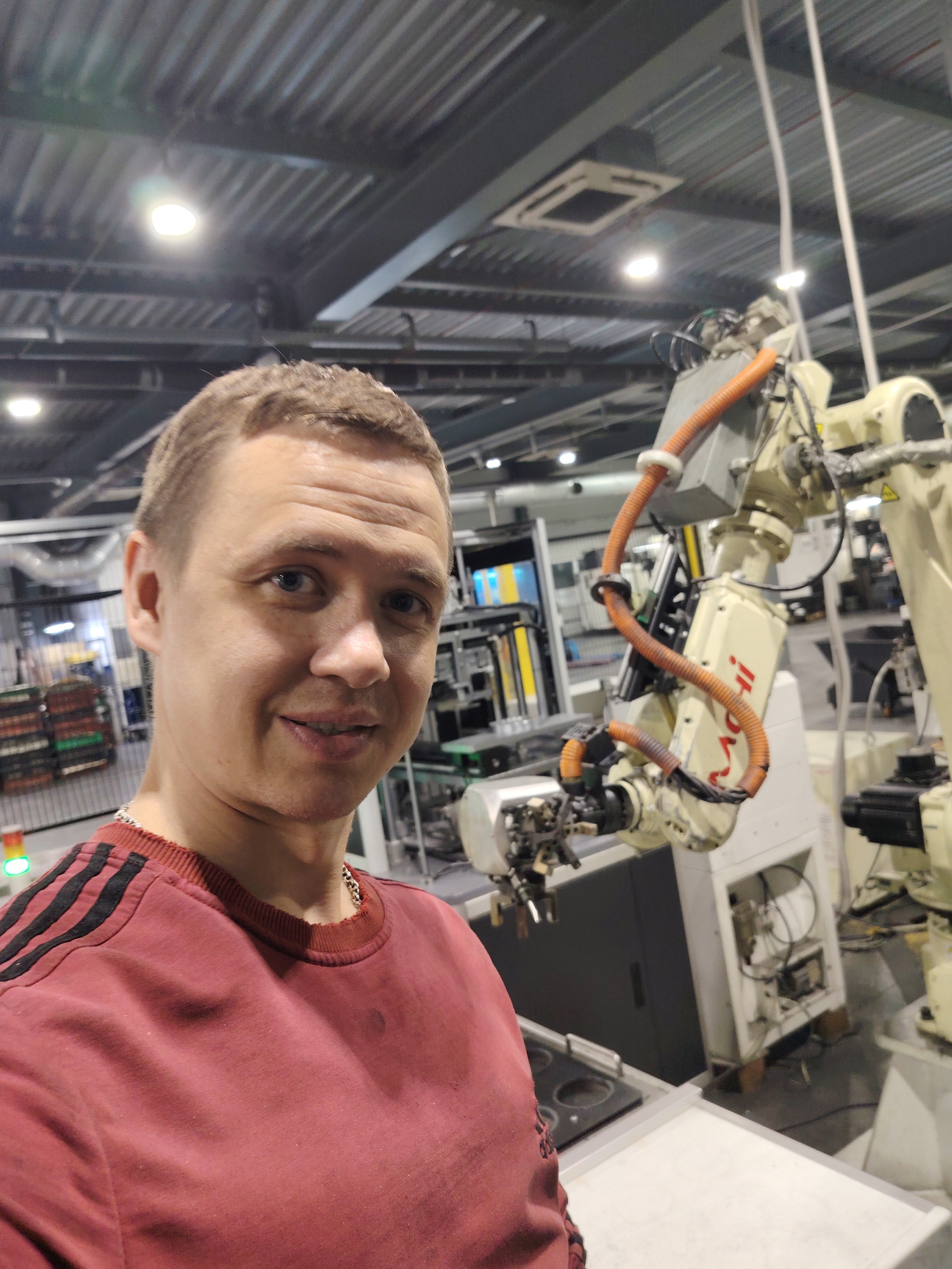 A fair-skinned man is taking a selfie in an industrial setting. He is wearing a maroon sweater with black stripes on the shoulders and a silver chain. Behind him stands a large robotic arm of a light cream color, with orange cables attached. The background shows a large warehouse-like space with visible ceiling beams, industrial lighting, and various pieces of machinery. There's a green light visible on the left side. The location cannot be determined to be Noviy, Russia based on the image alone.