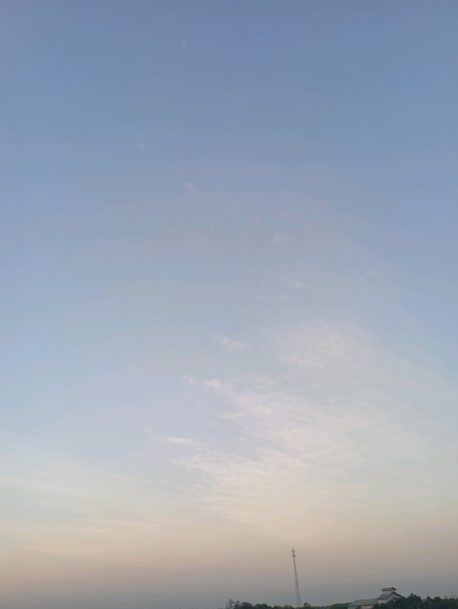The image shows a vast, pale blue sky with scattered wispy clouds, transitioning to a soft gradient of yellow and pink near the horizon. In the lower portion of the image, a faint silhouette of trees and some structures can be seen, suggesting a rural or semi-rural landscape. A tall, slender telecommunications tower stands prominently on the left side of the horizon line. To the right, a low-rise building with a peaked roof is visible amongst the trees. The scene appears to be captured at dawn or dusk. The location is Haurkolot, Indonesia.