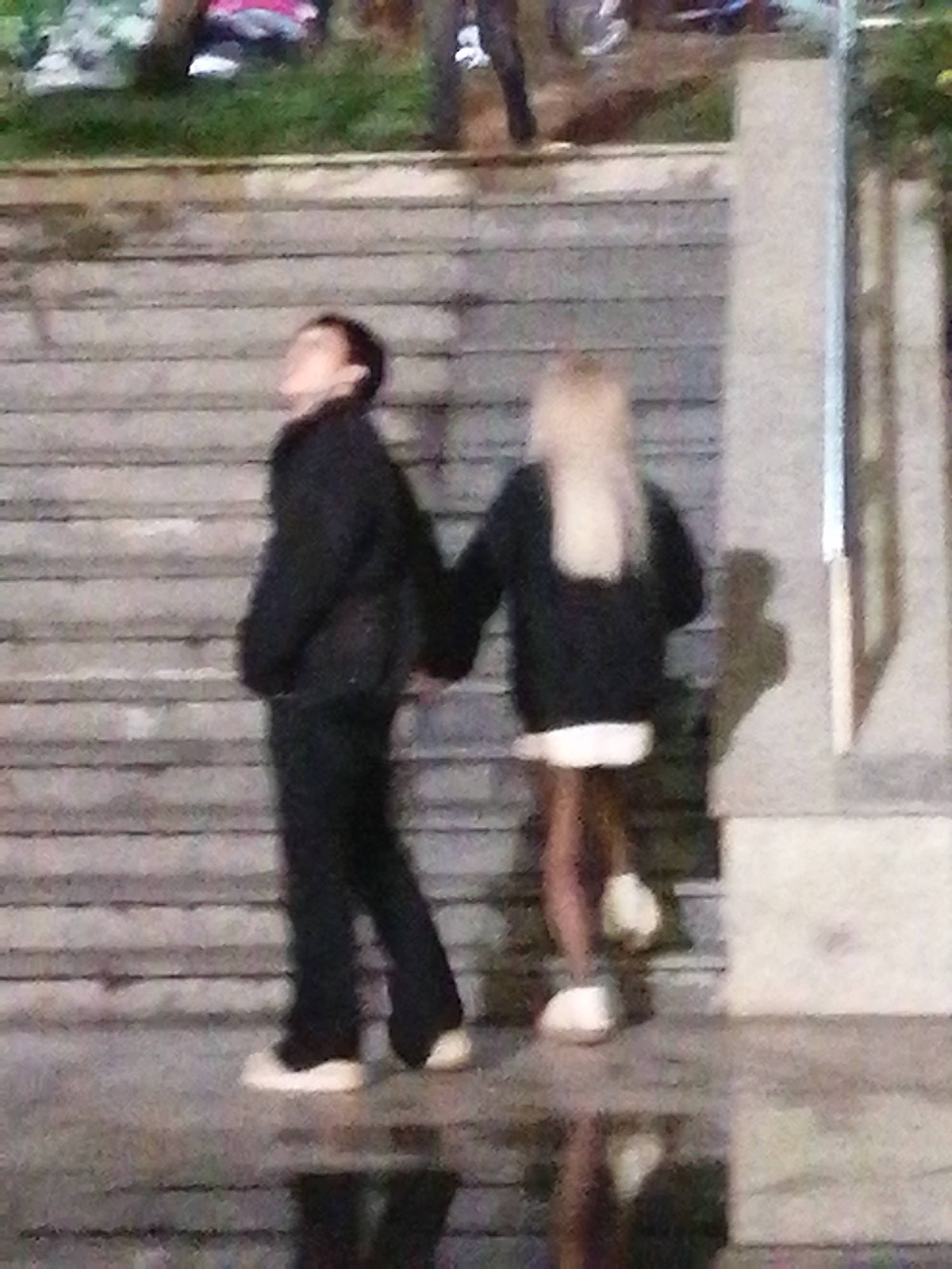 The image depicts two individuals standing on a set of wide, grey, concrete stairs. The person on the left is wearing a dark jacket and pants. The person on the right has long blonde hair and is wearing a dark jacket and a white skirt, with dark legs and white shoes. The background consists of what appears to be grassy areas and foliage. The lighting suggests it is either nighttime or dusk. The image quality is low, with significant pixelation and blurriness. The scene is located on a staircase, which could be found in various urban settings. Without additional context, it is impossible to definitively place the image in Da Lat, Vietnam.