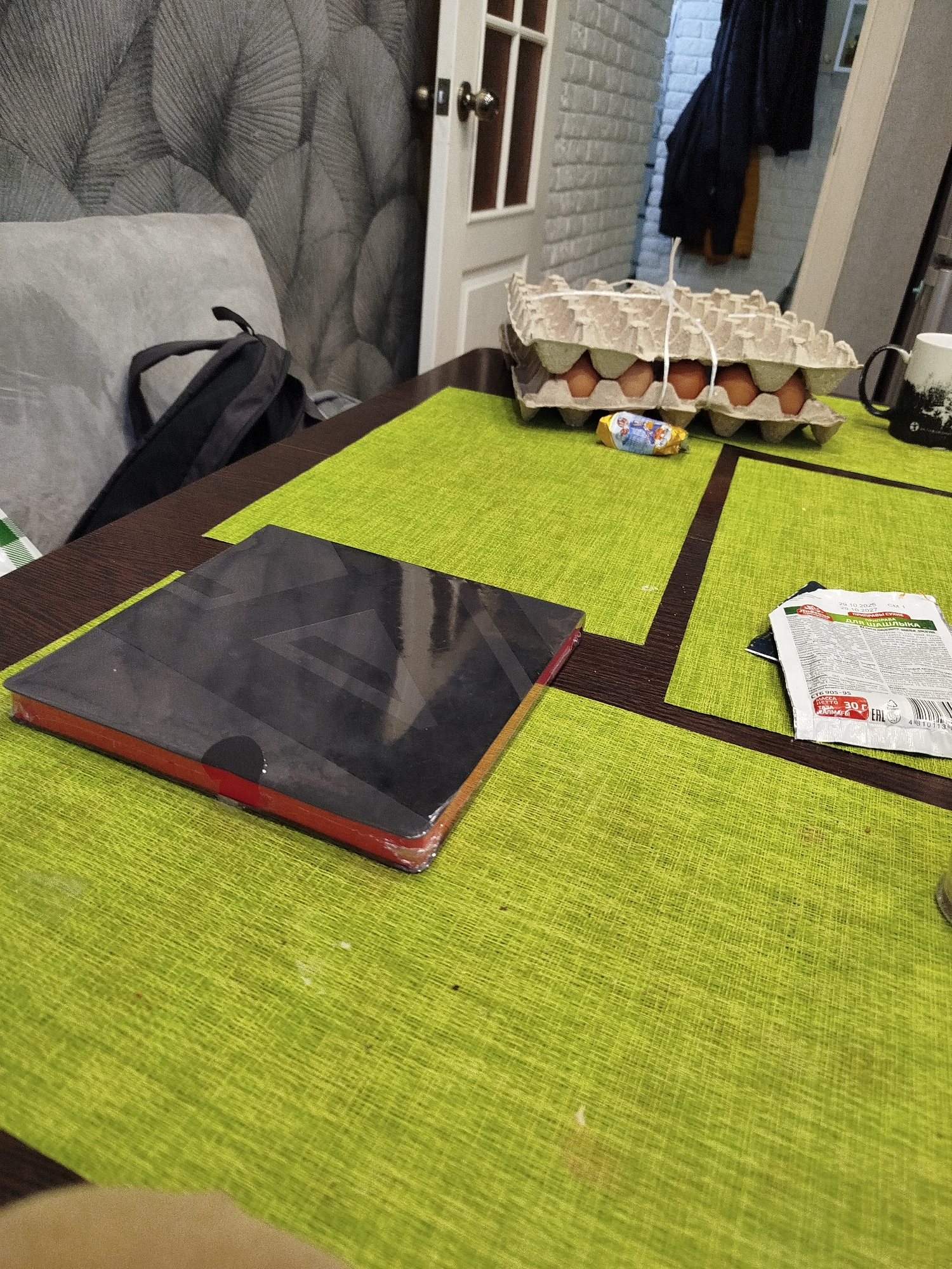 This image depicts a table set with a lime green placemat. On the placemat, there is a stack of black and red notebooks, an egg carton containing brown eggs, a small candy wrapper, and a packet of spice. In the background, a gray couch and a backpack are visible, along with a doorway and a wall with a textured pattern. The scene appears to be indoors, possibly in a residential setting, and suggests a casual environment. The presence of the placemat and the items on the table hint at a meal or a work-related activity.