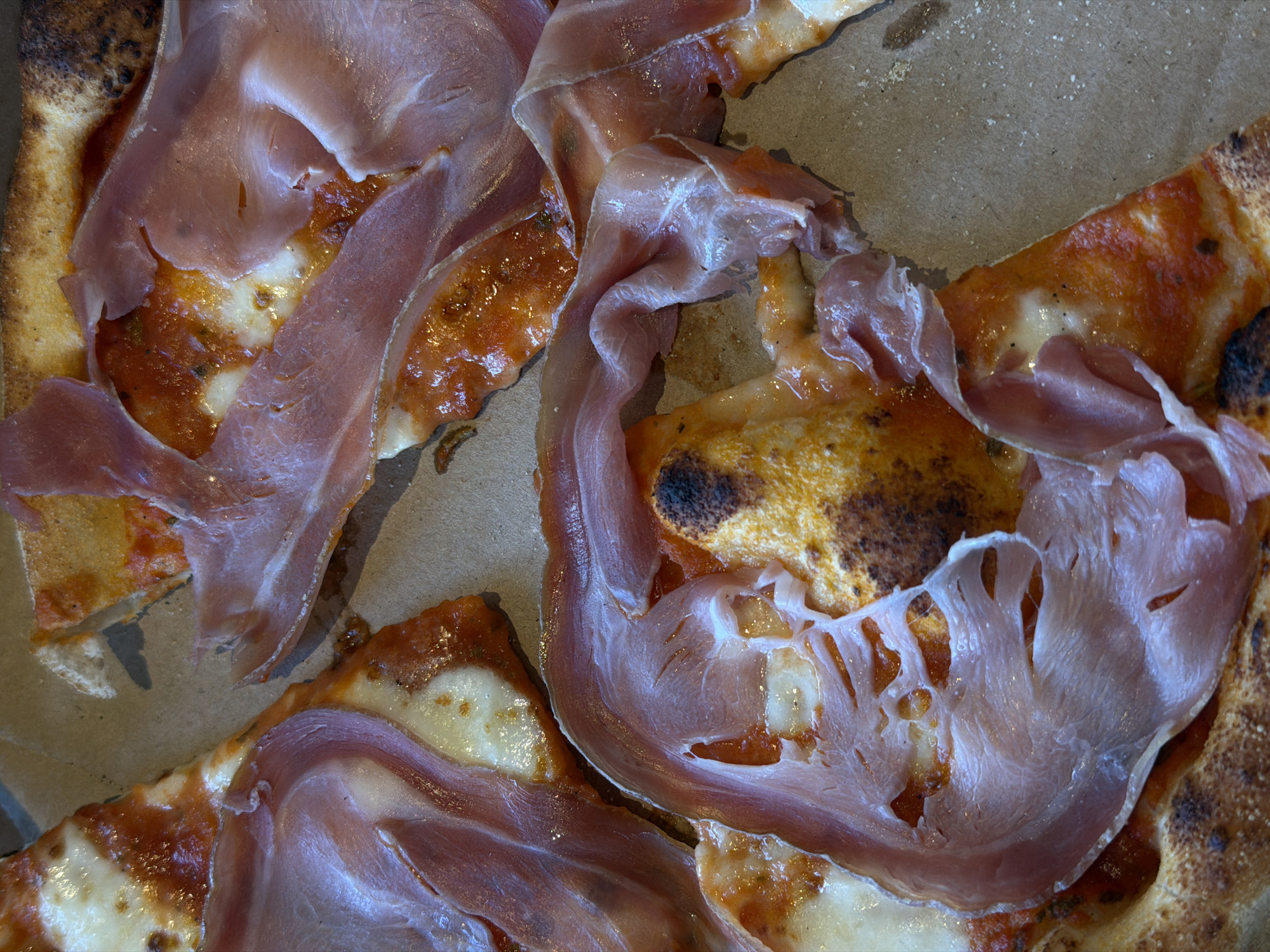 The image presents an overhead, close-up view of several slices of a Neapolitan-style pizza resting within a light brown, textured cardboard box. The pizza features a puffy, unevenly charred crust (cornicione), a vibrant reddish-orange tomato sauce base, and irregularly melted white cheese. Thinly sliced, translucent pink cured ham, specifically prosciutto, is draped over the pizza slices, covering portions of the cheese and sauce. One slice in the lower center shows significant charring on its central surface. The cardboard box displays slight texture and minimal food remnants. The image contains no visual elements or discernible features to identify its location as the city of Khimki, Russia, or any other specific geographic place. No individuals are visible.