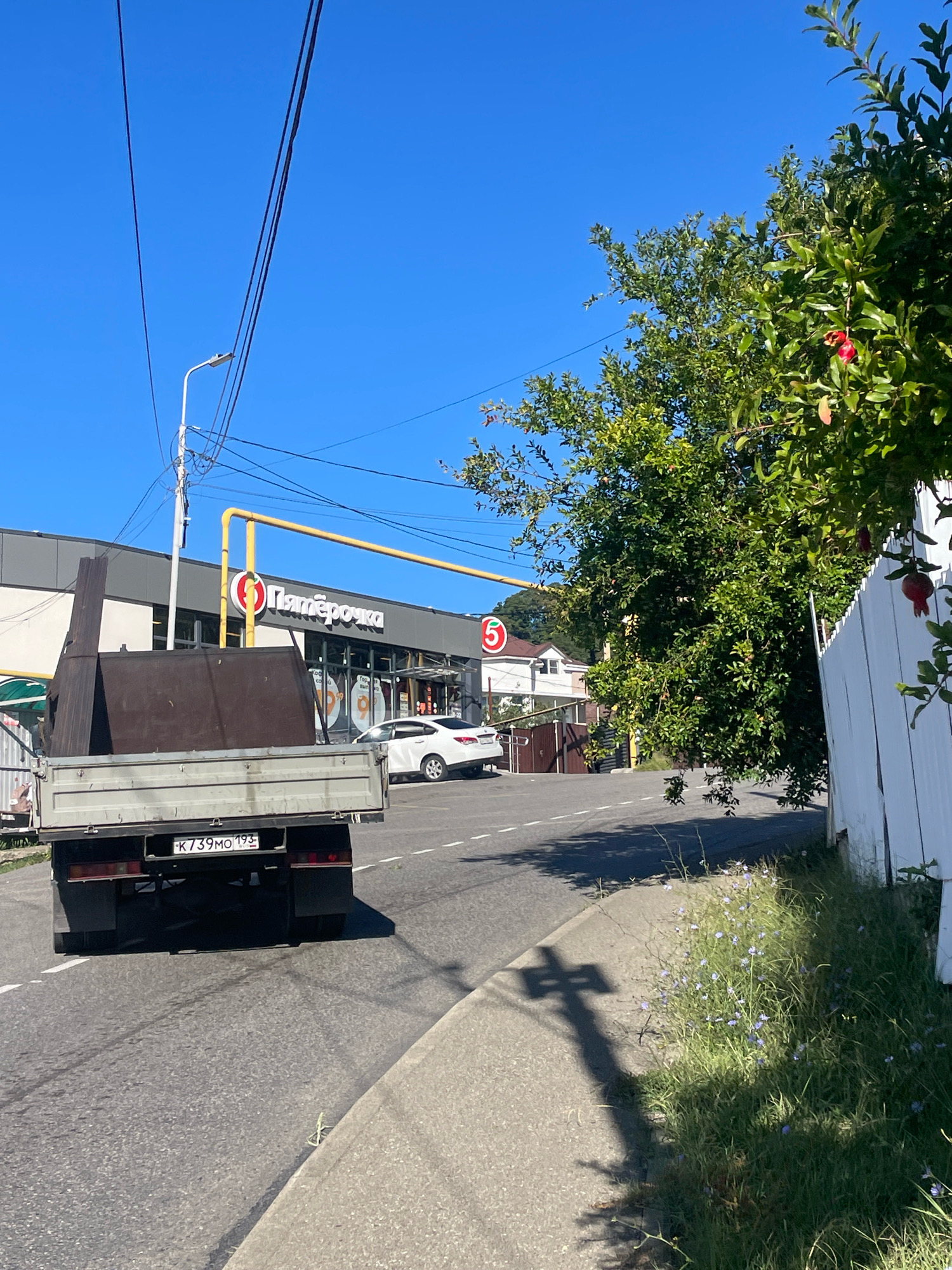 In Baranovka, Russia, a flatbed truck carrying large, dark metal panels is parked on a paved road. The truck's license plate reads "K739MO 193". To the left of the truck is a light-colored building with the "Пятёрочка" logo visible, indicating a store. Yellow gas pipes run diagonally across the scene in front of the building. A white car is parked in front of the store. A white picket fence runs along the right side of the image, bordering a grassy area with small purple wildflowers. A pomegranate tree with red fruit is visible in the background, partially obscuring a residential building. The sky is clear and blue.