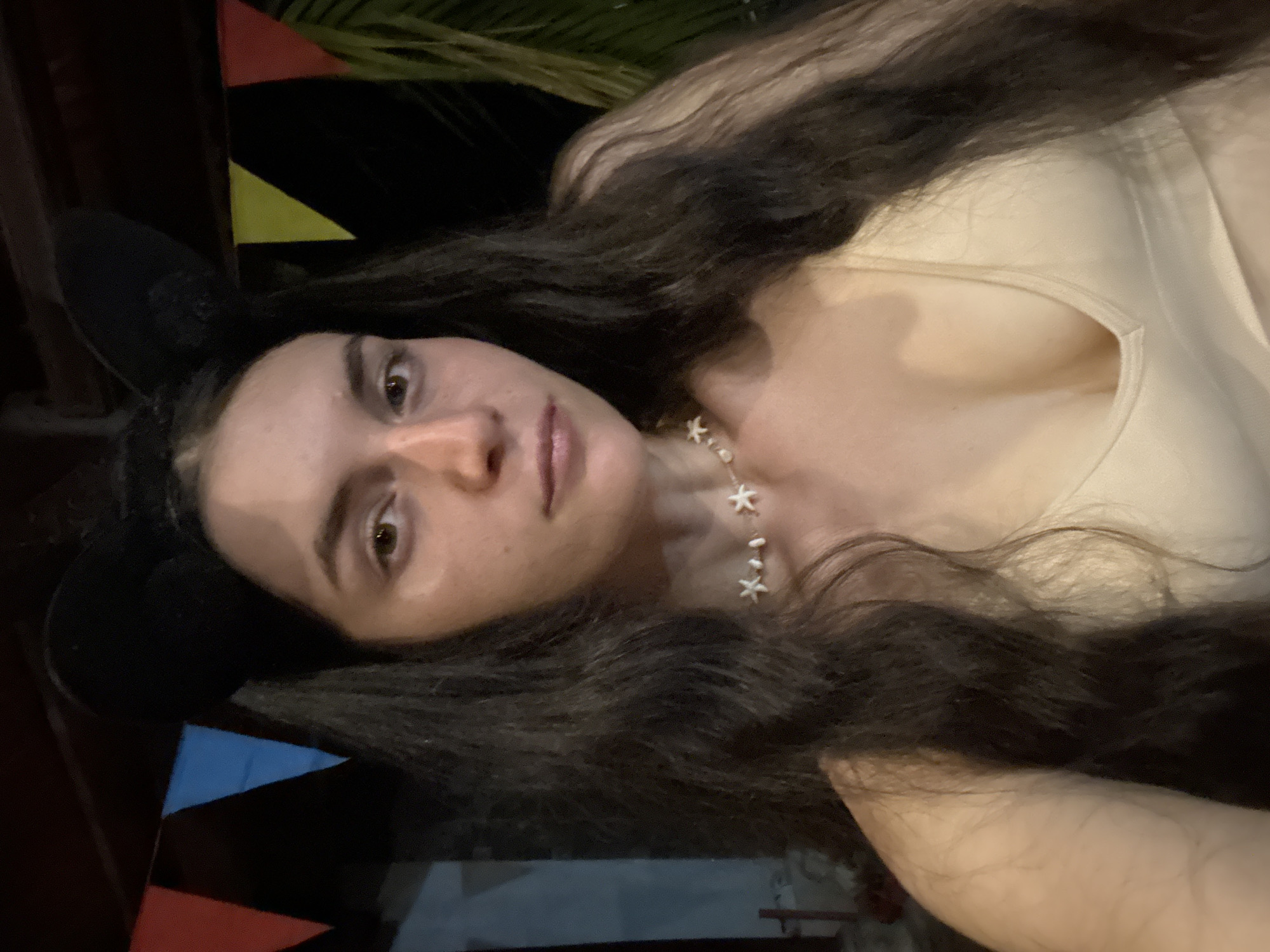A woman with long, dark, wavy hair and wearing black Mickey Mouse ears is looking at the camera. She is wearing a light-colored top and a starfish necklace. Red, yellow, and blue triangular pennants are visible in the background, along with some greenery. The location appears to be Mũi Né, Vietnam.