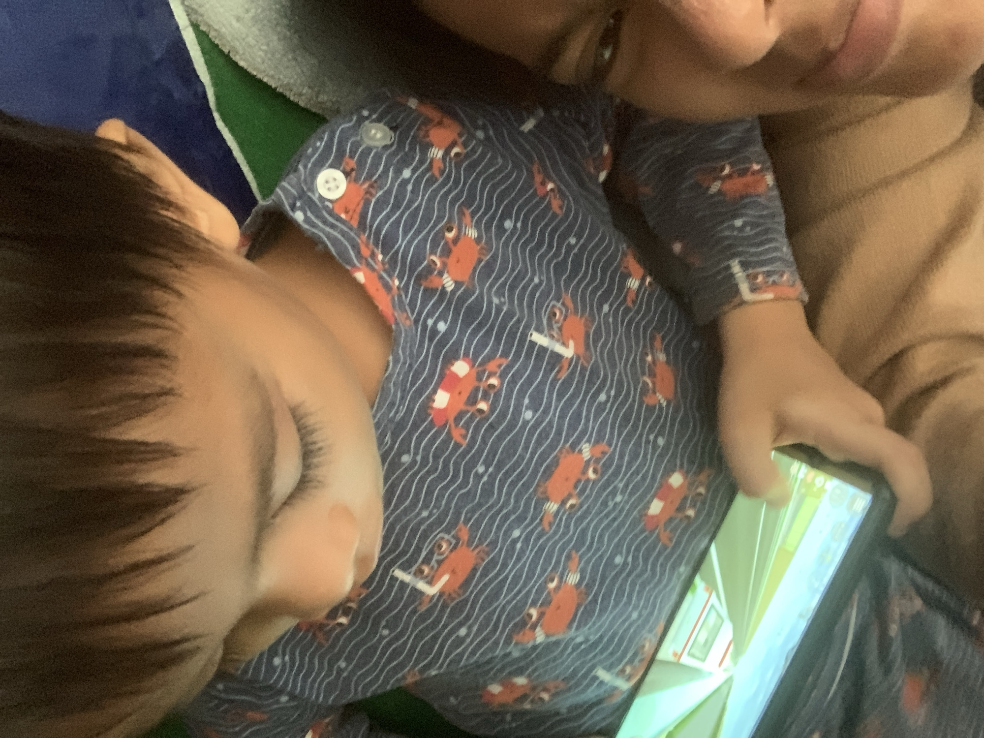 The media file shows a close-up, overhead view of two people, likely a child and an adult, in a relaxed setting. The child is wearing a dark blue pajama set with a pattern of orange crabs and white wavy lines. A white button is visible on the pajama top. The child's head and shoulder are in the foreground, with dark hair and closed eyes, suggesting they might be sleeping or resting.
The adult's face is partially visible in the upper right corner, with their lips close to the child's head. They are wearing a tan-colored garment. One of the adult's hands is visible holding a smartphone, which is displaying a bright, colorful screen with a scene that appears to be from a video game or animation. The screen shows vertical lines and what looks like a hallway or interior scene.
The background is composed of various soft textures, including a dark blue surface, a green and white striped fabric, and a gray, fuzzy material, possibly bedding or a blanket. The lighting appears to be indoor and soft, with no strong shadows, suggesting it could be daytime or evening. There is no visible text in the image. The overall impression is one of intimacy and comfort, with the adult sharing a quiet moment with the child, possibly while the child is resting or engaged with the phone. The location context provided is Sungai Meriam, Indonesia.
