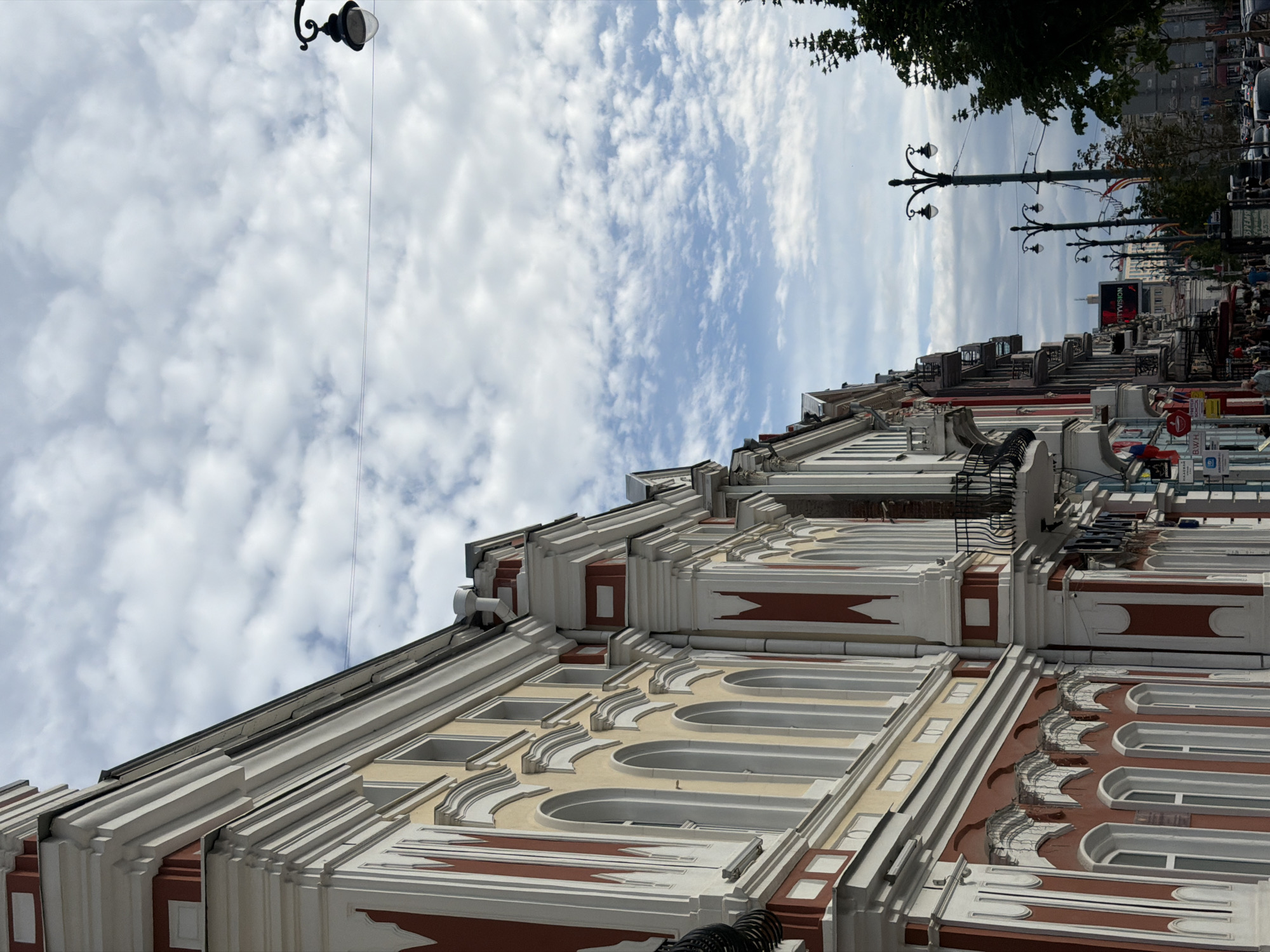 This is a photograph taken from street level, looking up at the facade of a row of buildings. The buildings are ornate, with red and cream-colored detailing, arched windows, and decorative cornices. Several balconies with black iron railings are visible. The sky above is partly cloudy with a blue sky peeking through. A few lampposts line the street, with one prominent lamppost in the foreground. Trees with green foliage are also present on the right side of the frame. There are some people walking on the sidewalk and a few cars visible in the distance. Signs and flags are attached to the buildings, indicating commercial establishments. The overall impression is of a historic urban street scene.
