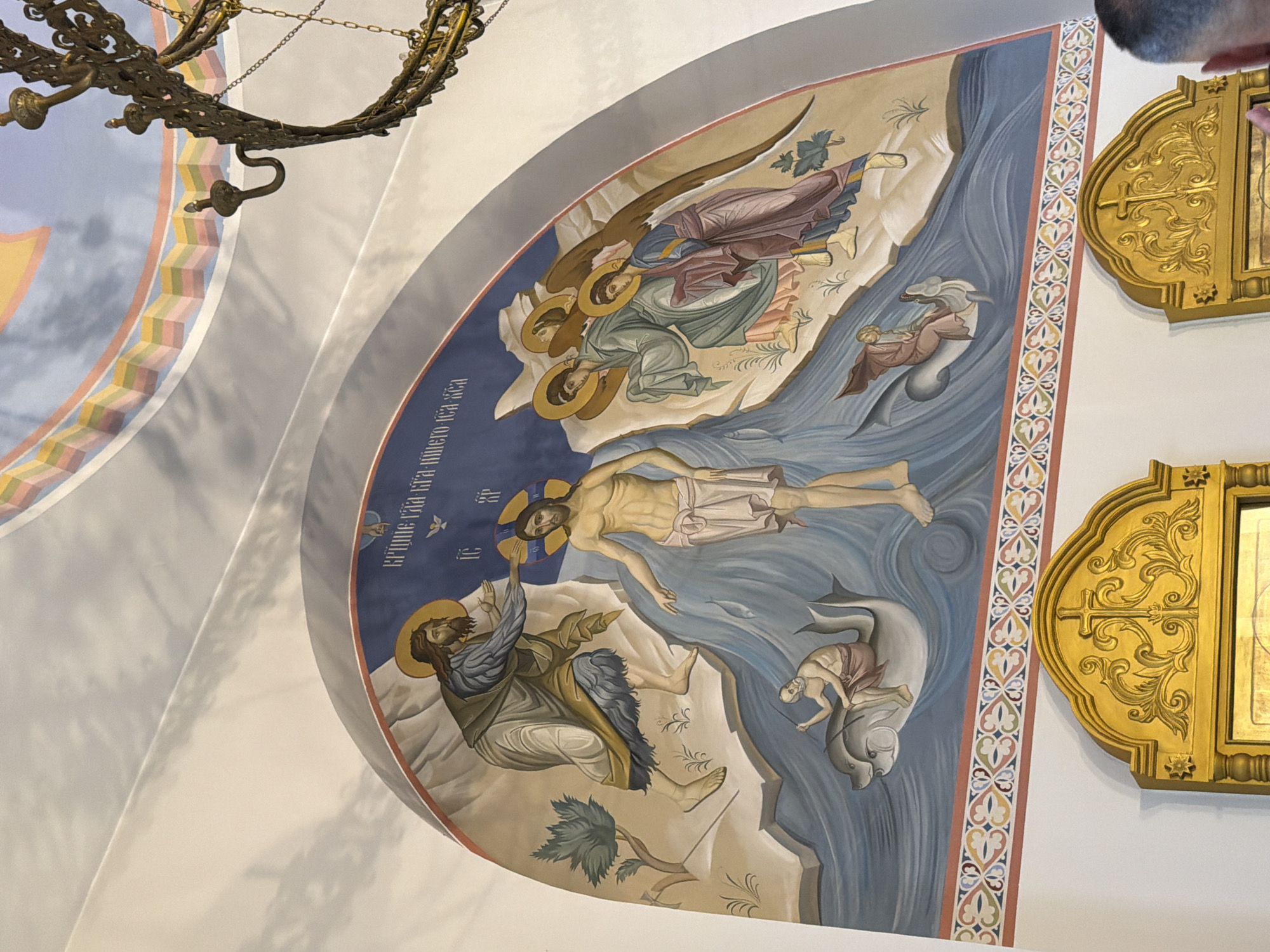 The image depicts a religious fresco adorning the interior of a building, likely an Orthodox Christian church, given the style of the artwork and the presence of icons below. The fresco, painted on a curved surface, illustrates a biblical scene. In the center, a figure identified as Jesus Christ, with a halo, is being baptized in a body of water. To his left, a bearded man, likely John the Baptist, with a halo, is pouring water on Jesus' head. Several angels with halos and wings are depicted on the right side of the scene, observing the baptism. The water is rendered with wavy lines, and there are fish visible within it. Small figures are seen riding marine creatures in the water below the main figures. The background features a rocky, stylized landscape and a blue sky with some white cloud-like elements. At the top of the fresco, there is text in an unfamiliar script, possibly Cyrillic. Below the fresco, ornate golden icon frames are visible, suggesting the presence of icons or religious paintings. The ceiling above the fresco has a decorative band of colorful geometric patterns and is painted with muted tones and shadows, suggesting it is also part of the architectural decoration. The lighting in the scene is bright, casting shadows that highlight the three-dimensional quality of the architectural elements and the artwork. The image does not contain information indicating its location in the city of Ko Samui District, Thailand.