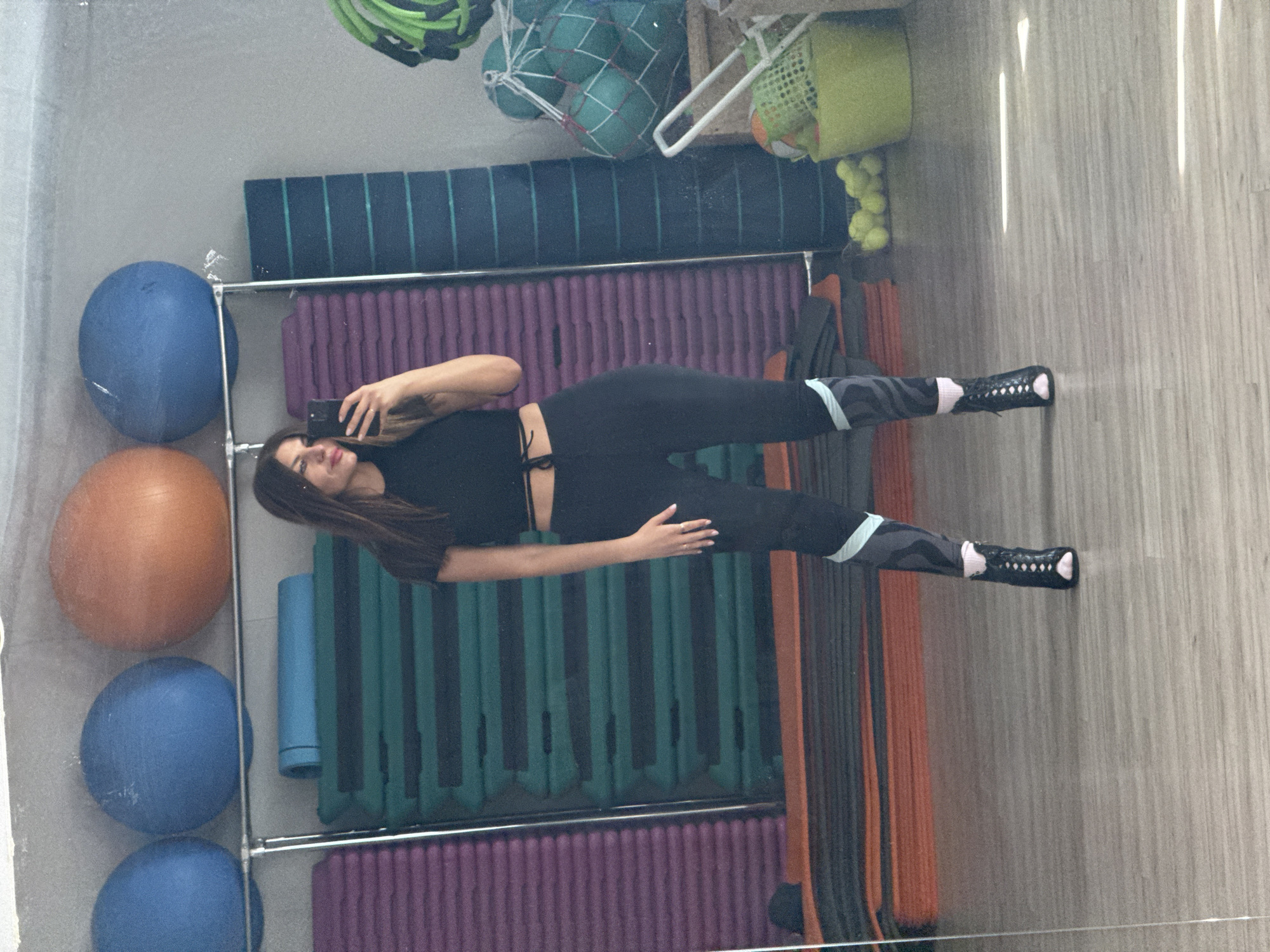 A mirrored photograph shows a woman in a black crop top and leggings posing with a mobile phone in what appears to be a gym or fitness studio. Behind her is gym equipment including exercise balls, step platforms, and mats. The location might be Chaikovsky, Russia.