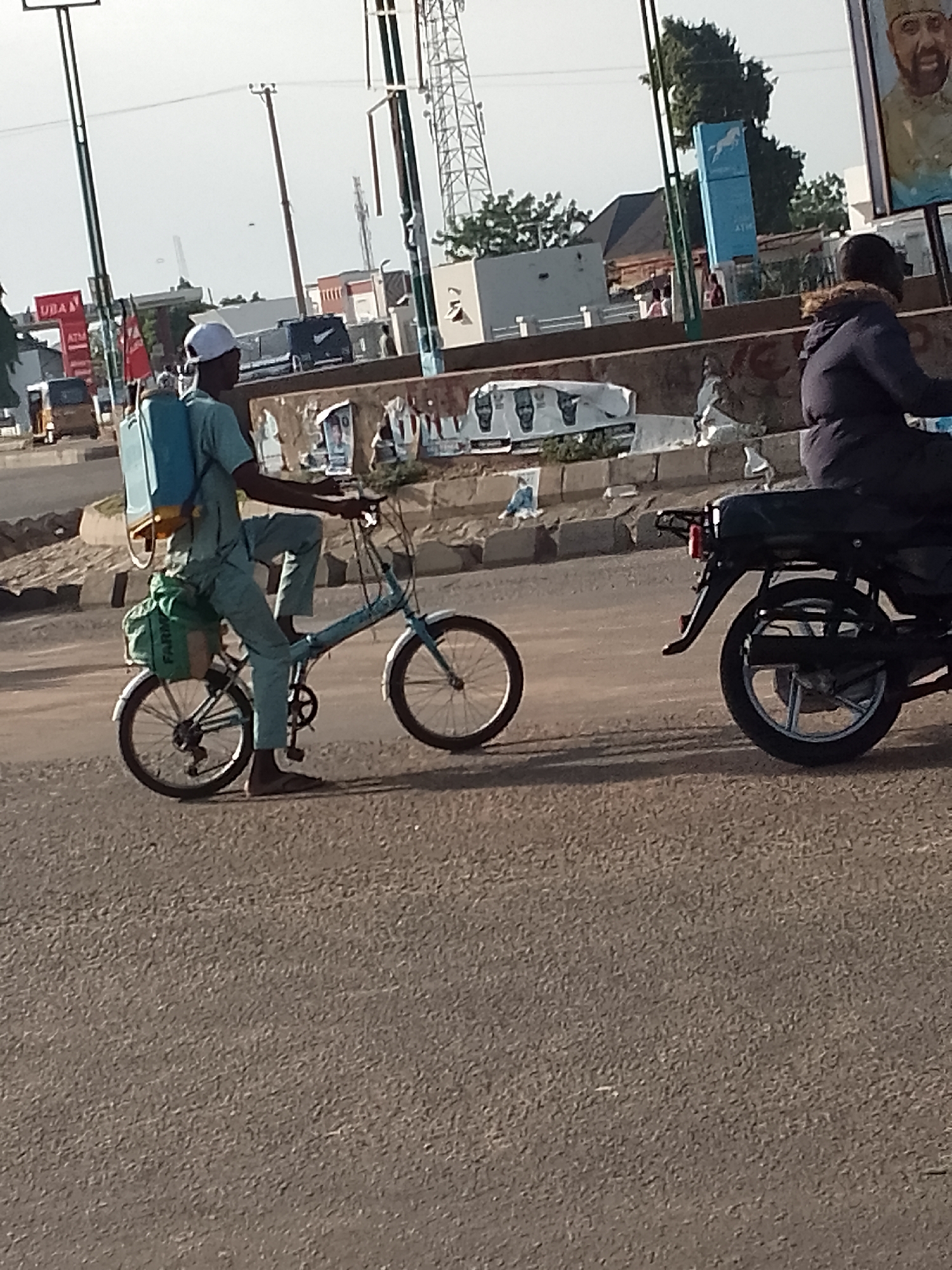 In Pantame, Nigeria, a man wearing a light blue outfit and a white cap cycles a blue foldable bicycle. He carries a large blue sprayer on his back and a green bag strapped to the rear of the bicycle. The man's right foot is resting on the pedal while his left foot is on the ground, indicating he is either starting or stopping. He is on a paved road with a sidewalk and roadside clutter in the background, including posters, buildings, and utility poles. To the right, a person on a black motorcycle is partially visible, also traveling on the road. The scene suggests an outdoor urban environment with typical street activity.