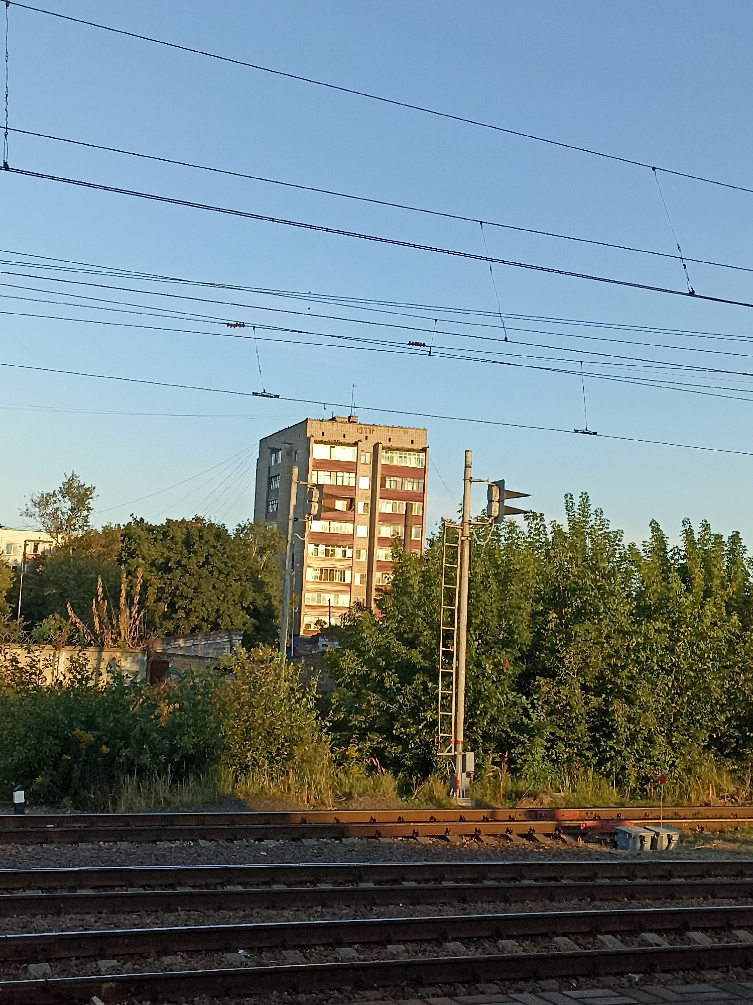 The image shows a view of railway tracks in the city of Chekhov, Russia, with a tall, multi-story apartment building in the background. Overhead power lines for the railway are visible against a clear blue sky. A railway signal with a ladder is also present near the building, surrounded by lush green trees. The foreground features multiple sets of parallel railway tracks, indicating an active railway area. The lighting suggests it is either early morning or late afternoon, with the sun casting shadows and highlighting the building and trees. There are no people visible in the image. The overall scene depicts an urban or semi-urban landscape with prominent railway infrastructure.