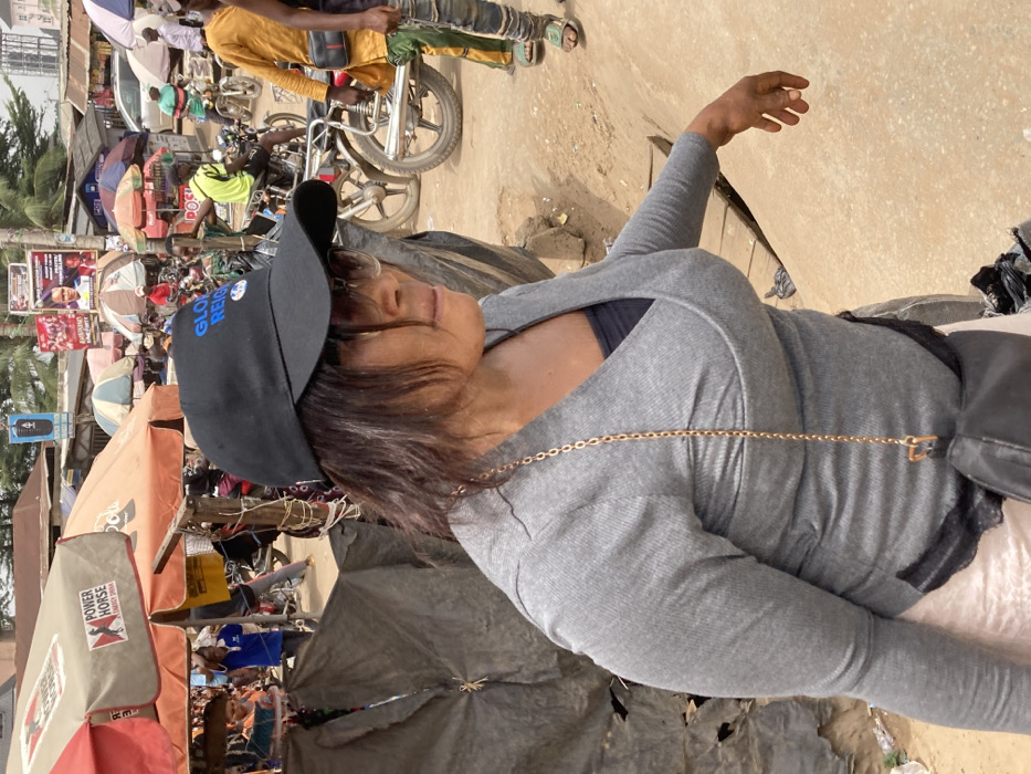 A forensic examination of the image reveals an outdoor market scene in Ura Nta Umuarandu, Nigeria.
The primary subject is a woman positioned in the foreground, facing right and slightly away from the camera. She wears a black baseball cap with "GLOBAL REIGN" in blue text, dark sunglasses, a grey long-sleeved ribbed wrap-style top over a black undershirt, and a light-colored skirt or lower garment. A black crossbody bag with a gold chain strap is visible across her torso. Her dark hair falls to her shoulders, and her left hand is partially visible, appearing to be in motion.
The background depicts a bustling market environment with a predominantly dirt ground and uneven terrain. Numerous individuals are scattered throughout the scene, some walking or standing near market stalls. Multiple motorcycles are parked or being operated. Market infrastructure includes several awnings and umbrellas, one prominently displaying "POWER HORSE ENERGY DRINK" branding. Dark tarpaulin sheets form makeshift shelters or stalls. In the midground and background, various buildings are visible, alongside multiple large posters and banners affixed to poles, some appearing to be political campaign advertisements (e.g., "PRINCE," "EMMANUEL ANYANWU"). Overhead utility lines are present. The lighting suggests a bright, sunny daytime setting.