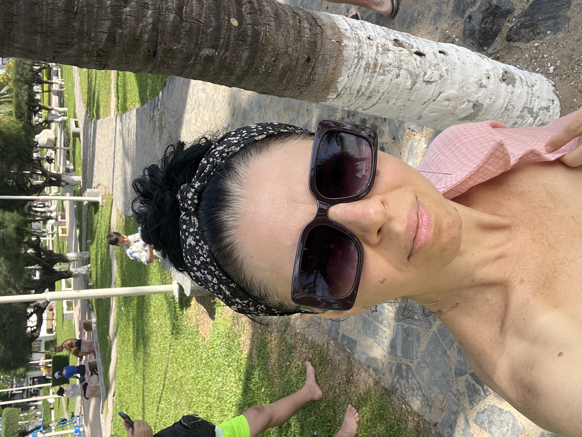 A woman wearing a pink top and a floral headband is taking a selfie in a park. She has large, dark sunglasses on. A palm tree trunk is visible to her right. In the background, a paved path winds through a grassy area with trees and benches. Several other people are scattered throughout the park, including a man and woman sitting on a bench and a child looking at a phone. The scene appears to be a public park in Nha Trang, Vietnam, possibly near a beach given the palm trees and casual attire of the individuals.
