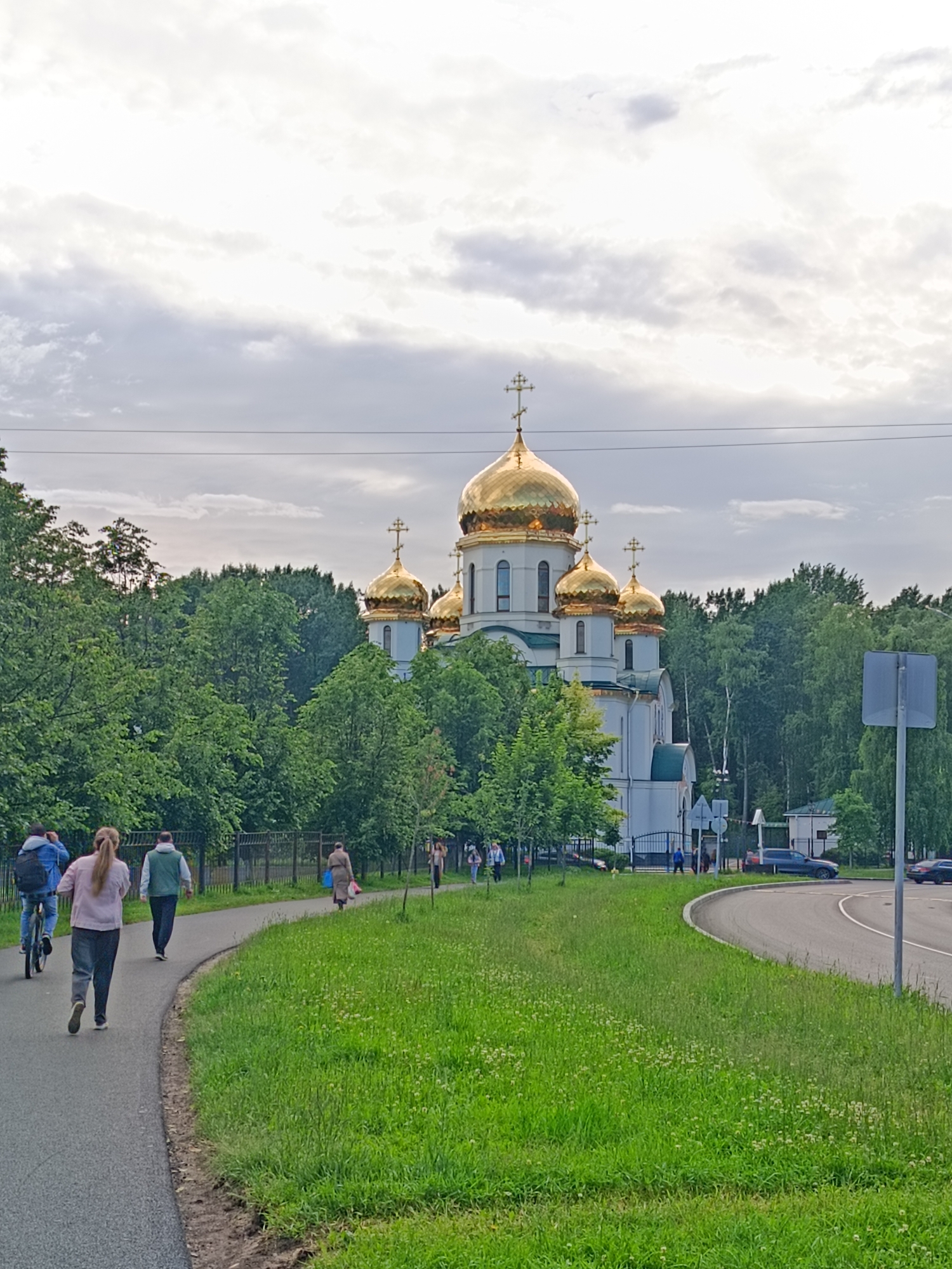 The image shows a church with golden domes in the city of Saint Petersburg, Russia, on a cloudy day. In the foreground, a paved path curves to the right, with a grassy area to the right of the path. Several people are walking and cycling on the path. Behind the path and grassy area is a fence and then trees, with the church visible behind the trees. The church is white with multiple golden domes and crosses. A road is visible to the right of the church, with a few cars driving on it. The sky is overcast with scattered clouds.
