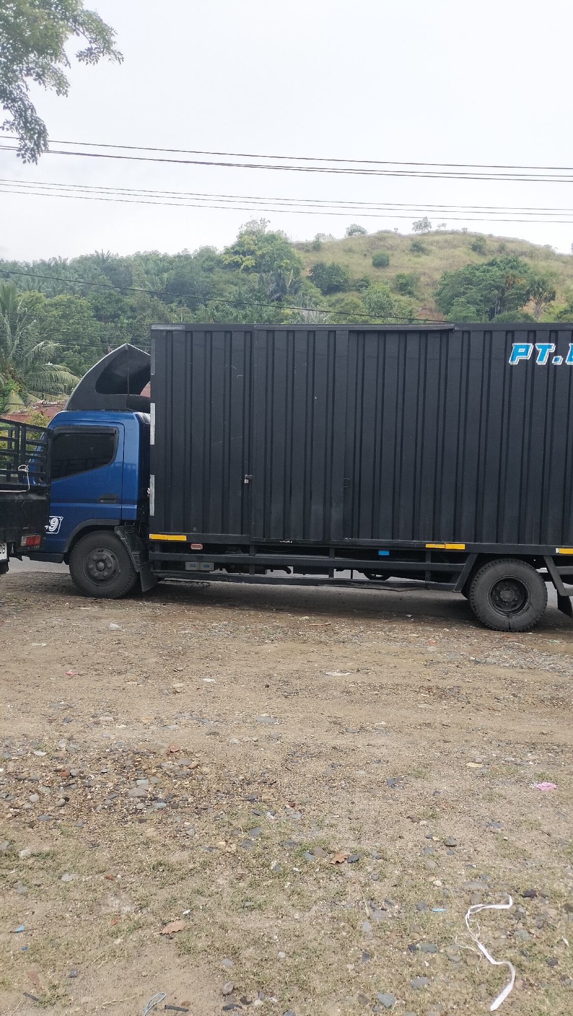 A blue and black truck is parked on a dirt and gravel lot. The truck has a black cargo container attached to its chassis. The container has the partial text "PT.L" in blue lettering on its side. The truck is a medium-duty truck with a single cab. The truck appears to be parked on the side of a road, with a wooded hillside in the background. The sky is overcast. The location is Lhokseumawe, Indonesia.