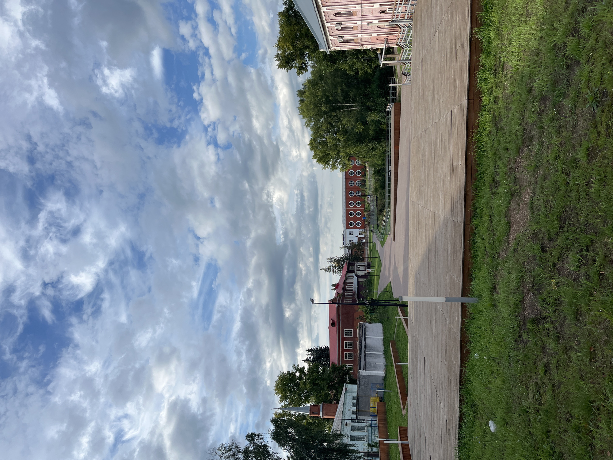 The image depicts an outdoor public space with a large wooden deck in the foreground, covered in green grass and some patchy soil. Beyond the deck, a paved pathway curves through the scene, lined with trees and buildings. In the background, several structures are visible, including a red-roofed building with white-trimmed windows, a pale blue building with a tall, slender spire, and a pink building with arched windows. A building with a repeating pattern of circular windows is also present. The sky is filled with a mix of white and gray clouds. The scene appears to be a park or a public square in the city of Blagoveshchensk, Russia, likely a renovated or newly developed area, given the modern wooden decking and paved pathways. No people are visible in the image.