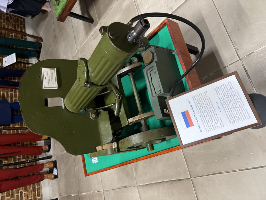 A green Russian Maxim Machine Gun, model 1910, sits on a wooden stand. A sign with the Russian flag indicates the gun's name. The sign describes it as a heavy machine gun used by the Imperial Russian Army during World War I and the Red Army during World War II. It was adopted in 1910 and chambered for the 7.62x54mmR cartridge. The M1910 was mounted on a wheeled mount with a gun shield. It was replaced in Soviet service in 1941.