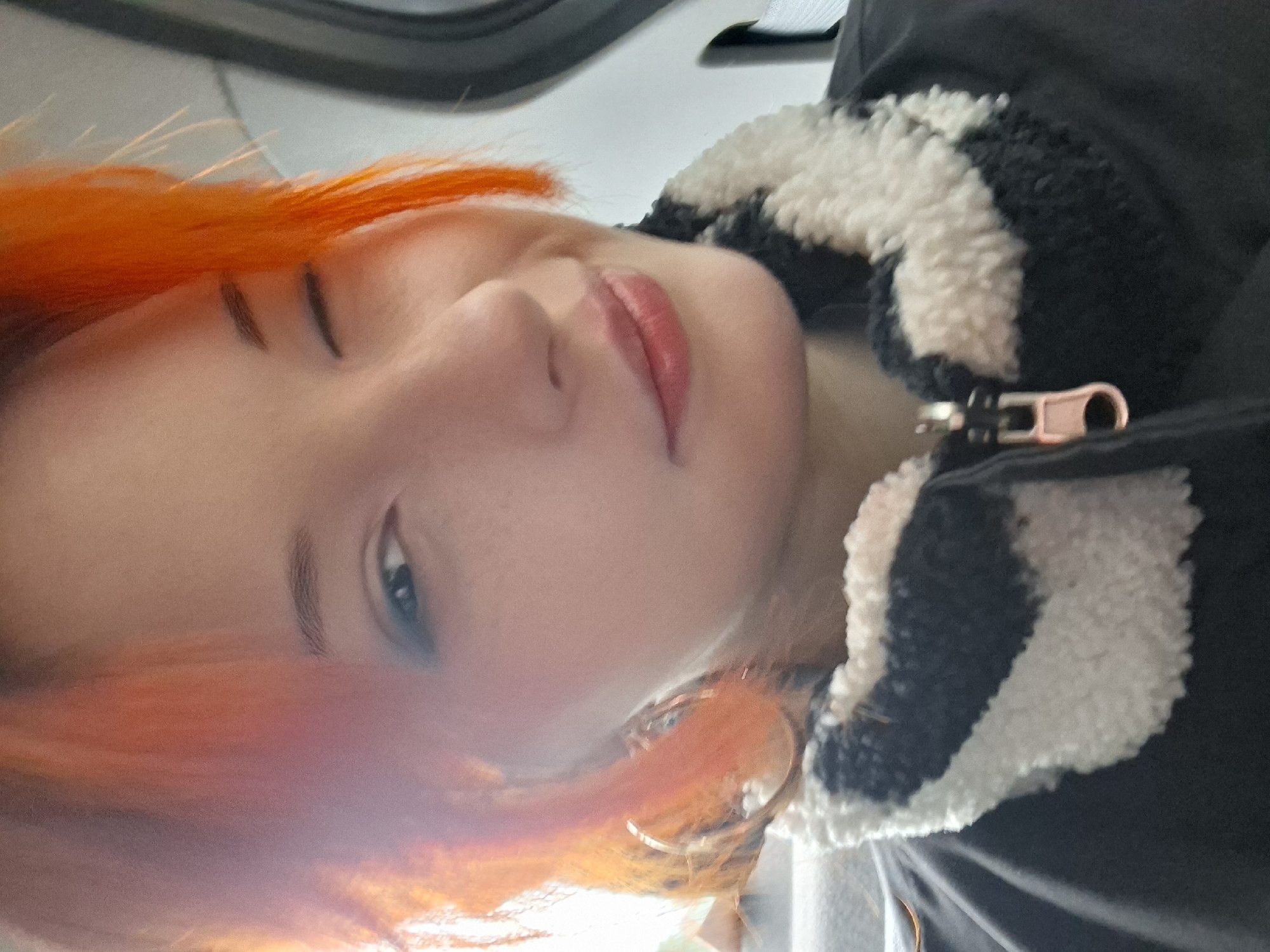 A close-up, slightly out-of-focus selfie of a person with orange hair. The person is wearing a dark jacket with a white and black striped fleece collar. The person's face is angled towards the camera, showing a single eye, eyebrow, nose, and lips with reddish lipstick. The location is not discernible from the image.