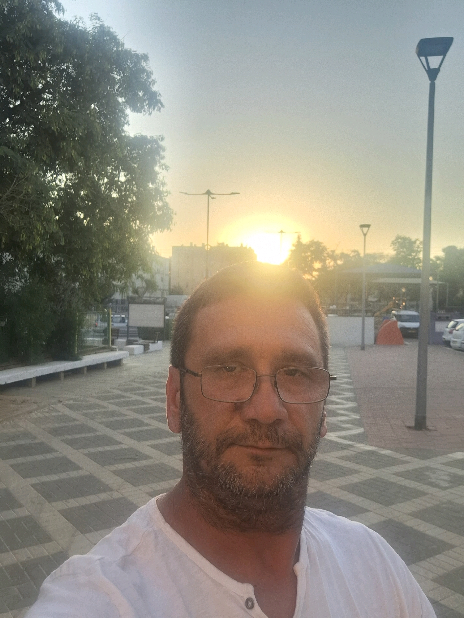 A man with glasses and a beard, wearing a white t-shirt, is taking a selfie outdoors at sunset in Yerocham, Israel. The sun is setting in the background, casting a warm glow. In the foreground, the man's face and upper body are visible. Behind him, there is a paved walkway with a geometric pattern, trees, buildings, and streetlights. The overall scene depicts a casual moment in an urban setting during dusk.