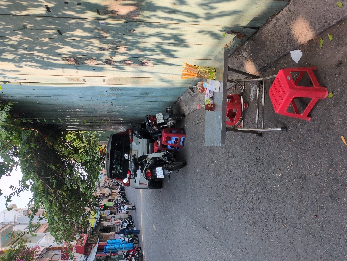 The scene depicts a narrow urban street in Ho Chi Minh City, Vietnam, under bright daylight with dappled sunlight creating strong shadows.
In the immediate foreground, a small, rectangular stainless-steel folding table is positioned on the roadside against a faded light-blue corrugated metal wall. On the table, a clear glass vessel holds numerous yellow chopsticks, alongside a small red-capped condiment bottle, a tissue dispenser, and three small white ceramic dishes. Two red plastic stools are present, one directly in front of the table and another partially visible tucked underneath. On the dark asphalt pavement surrounding the table, a crumpled white tissue, several scattered green leaves, and a small reddish object are visible.
Behind the table, two motorbikes are parked; one is dark-colored and partially obscures a blue plastic stool. Further down the street, a white SUV is parked facing away from the camera, partially on the asphalt and partially near the corrugated wall. A line of additional motorbikes is parked along the left side of the asphalt road.
The left side of the street is lined with multi-story buildings, characteristic of urban Vietnamese architecture, featuring various pastel colors, balconies, and ground-floor storefronts. One shop displays several large, rolled blue items. Several individuals are visible in the mid-distance: one person stands near the blue items, and another is seen riding a motorbike further down the street. A canopy of dense green tree foliage extends over the street from the left, casting shadows onto the corrugated wall. The corrugated wall itself shows signs of wear, including faded paint and darker stains.