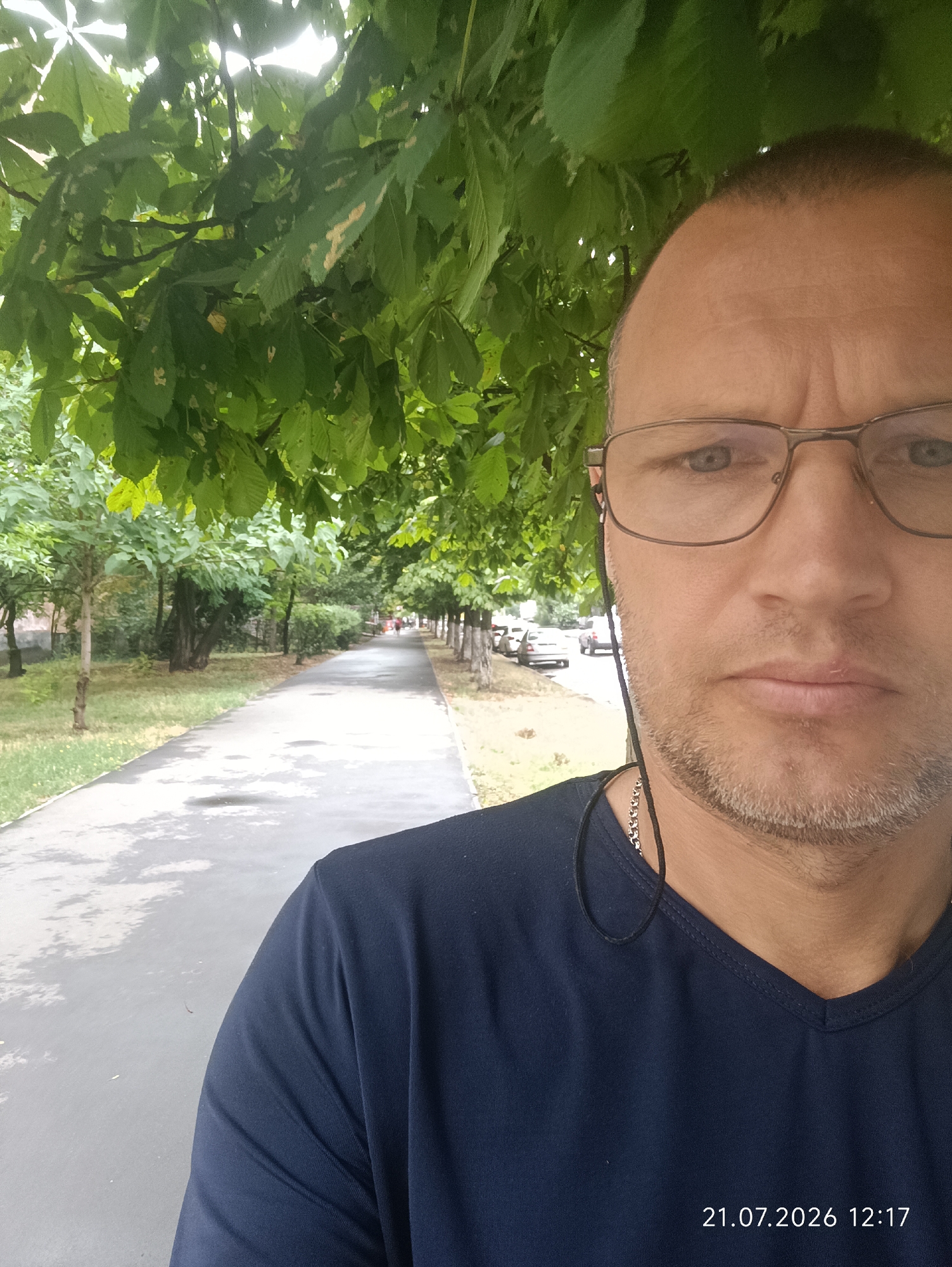 The image is a selfie taken outdoors, with a man's face filling the right side of the frame. He is wearing glasses and a dark blue V-neck t-shirt, with a silver chain visible around his neck. Earbuds are tucked behind his ears. Behind him, a tree-lined path stretches into the distance, with cars parked along the side of the road. The path appears wet, suggesting it may have recently rained. The foliage above the man is lush and green, indicating a summer day. A timestamp in the lower right corner reads "21.07.2026 12:17". The scene is consistent with a park or residential street in Odesa, Ukraine.